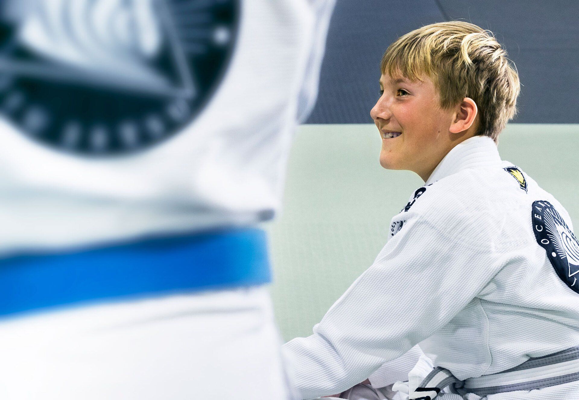 Young person in a white gi smiles during a martial arts class; blurred blue belt and logo in the background.