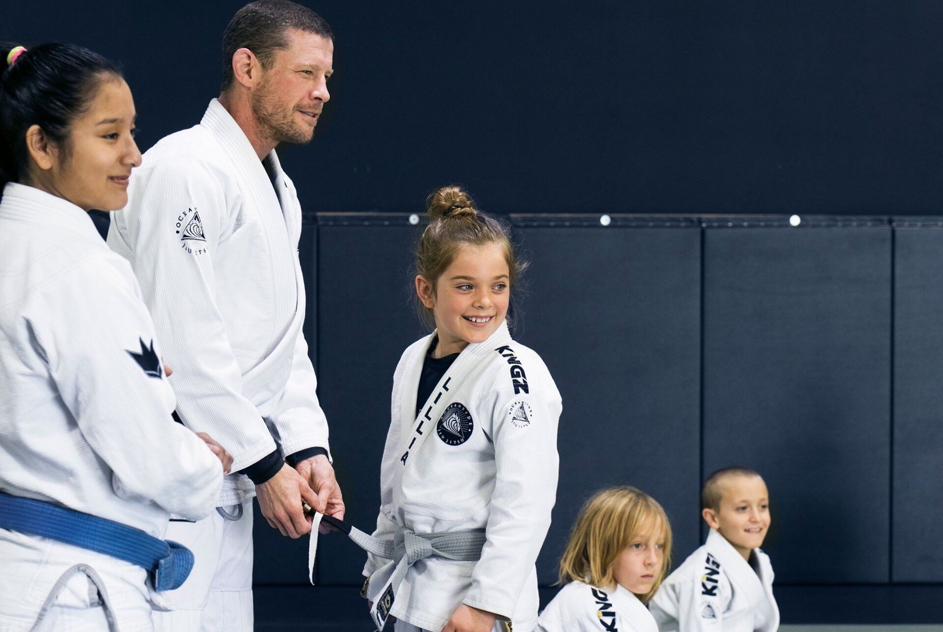 People in white martial arts uniforms, smiling, with a black mat background.