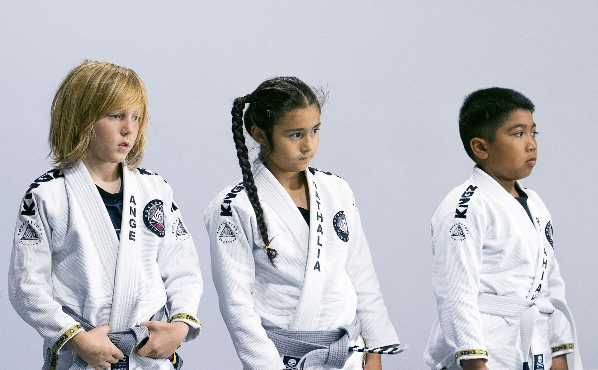 Three children in white martial arts uniforms stand indoors, looking forward.
