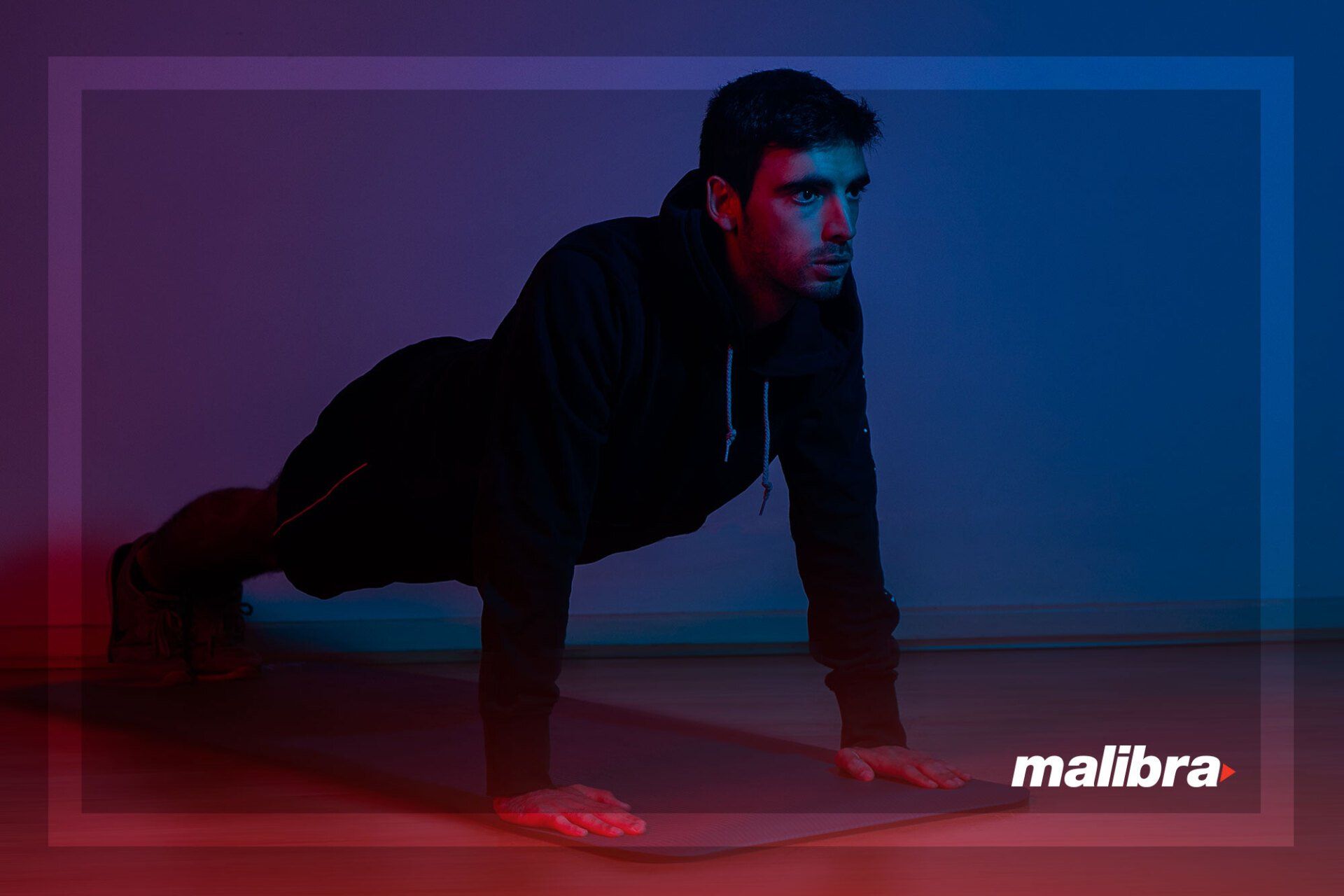 Man in a black hoodie doing push-ups on a mat, indoors, lit with blue and red light.