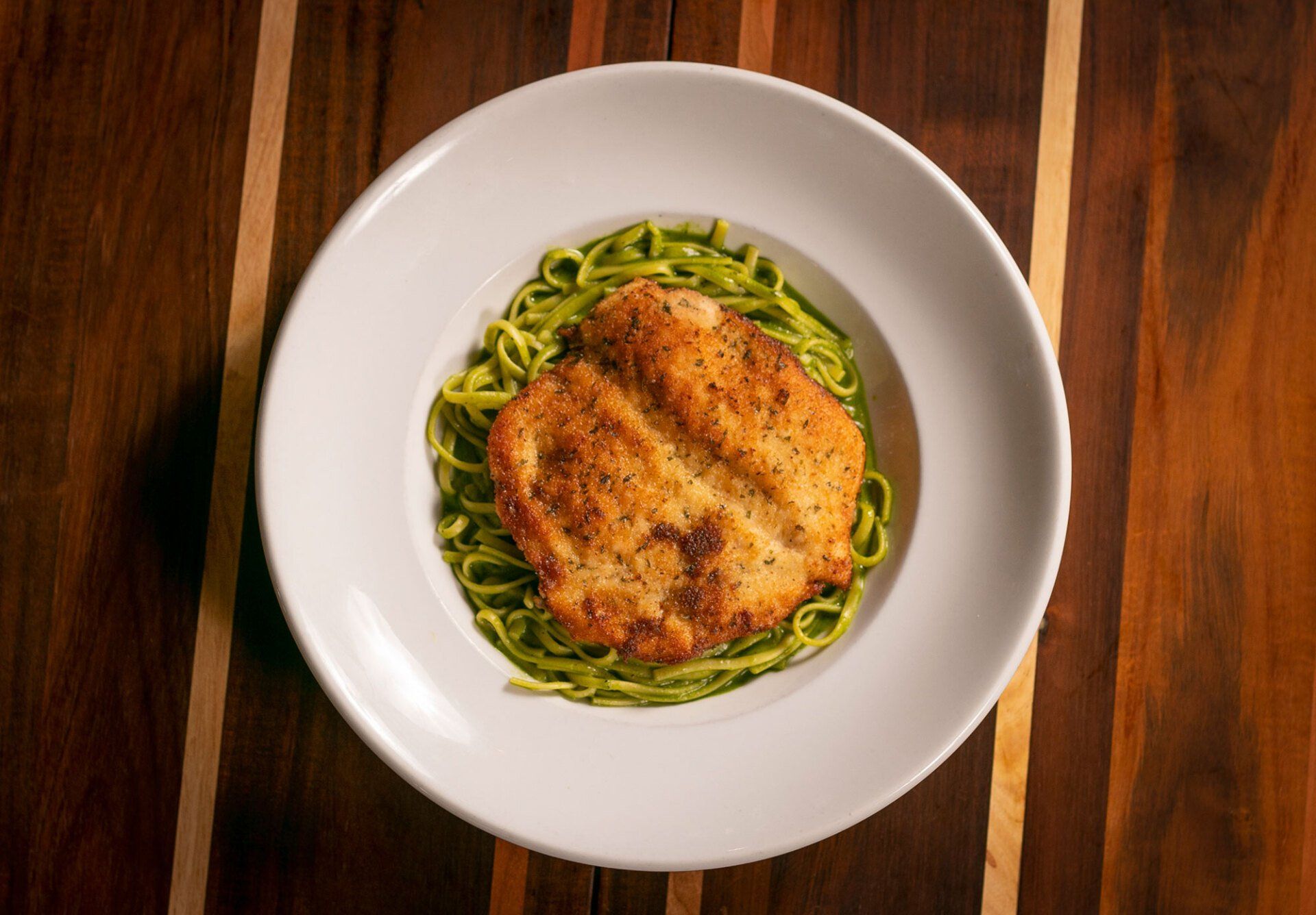 Chicken breast on green pasta in a white bowl, on a wood table.