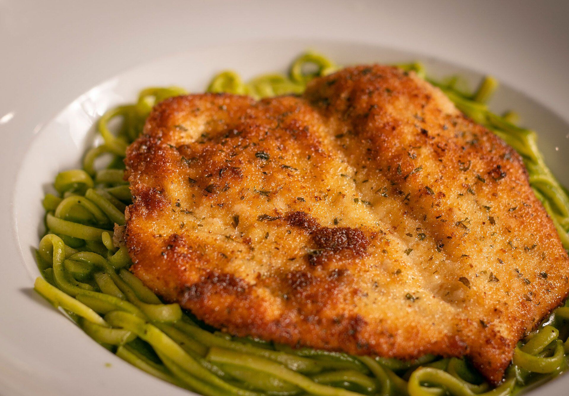 Breaded chicken cutlet over green pasta in a white bowl.