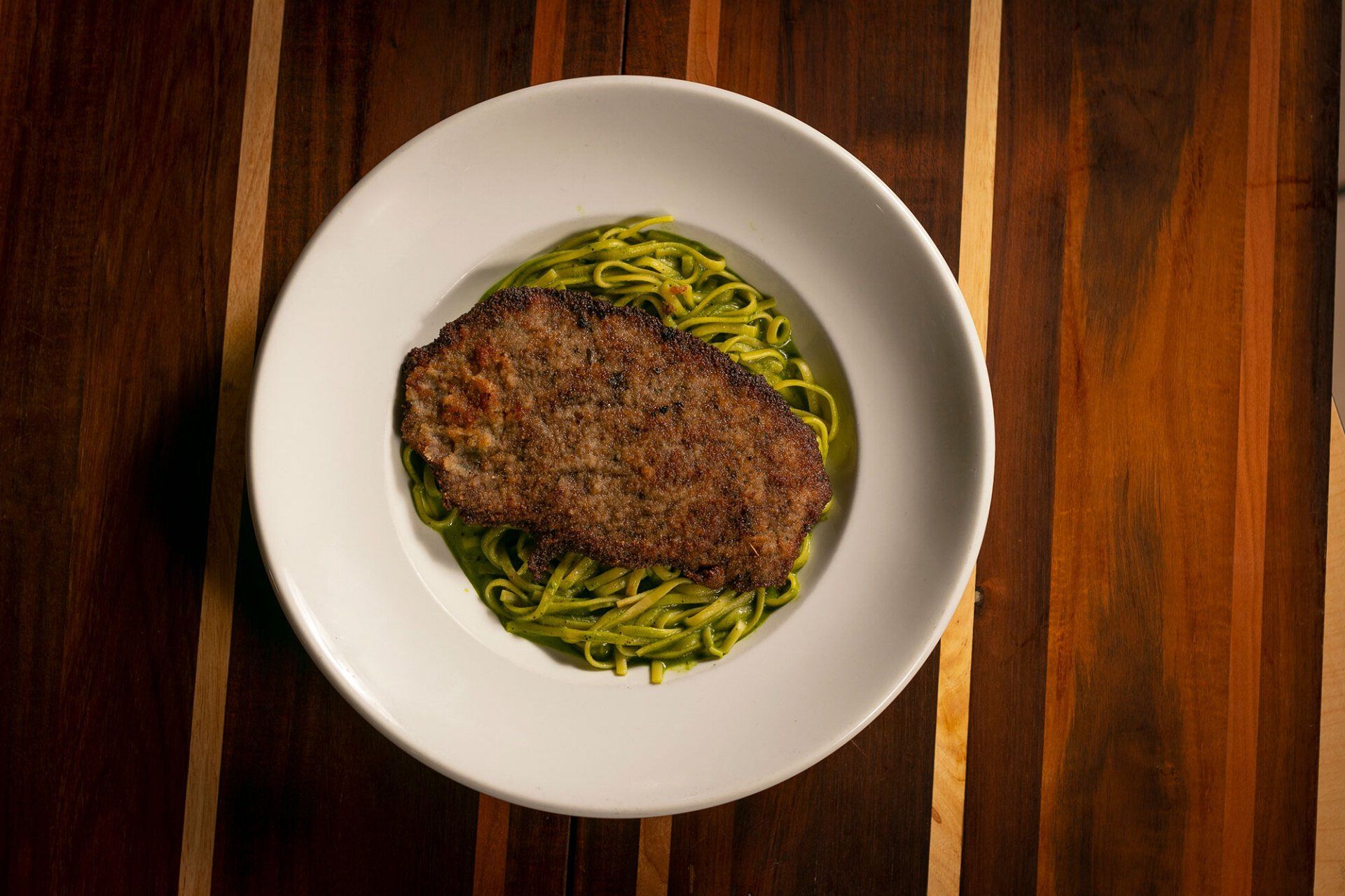 A steak atop green pasta in a white bowl, set on a wooden surface with brown stripes.