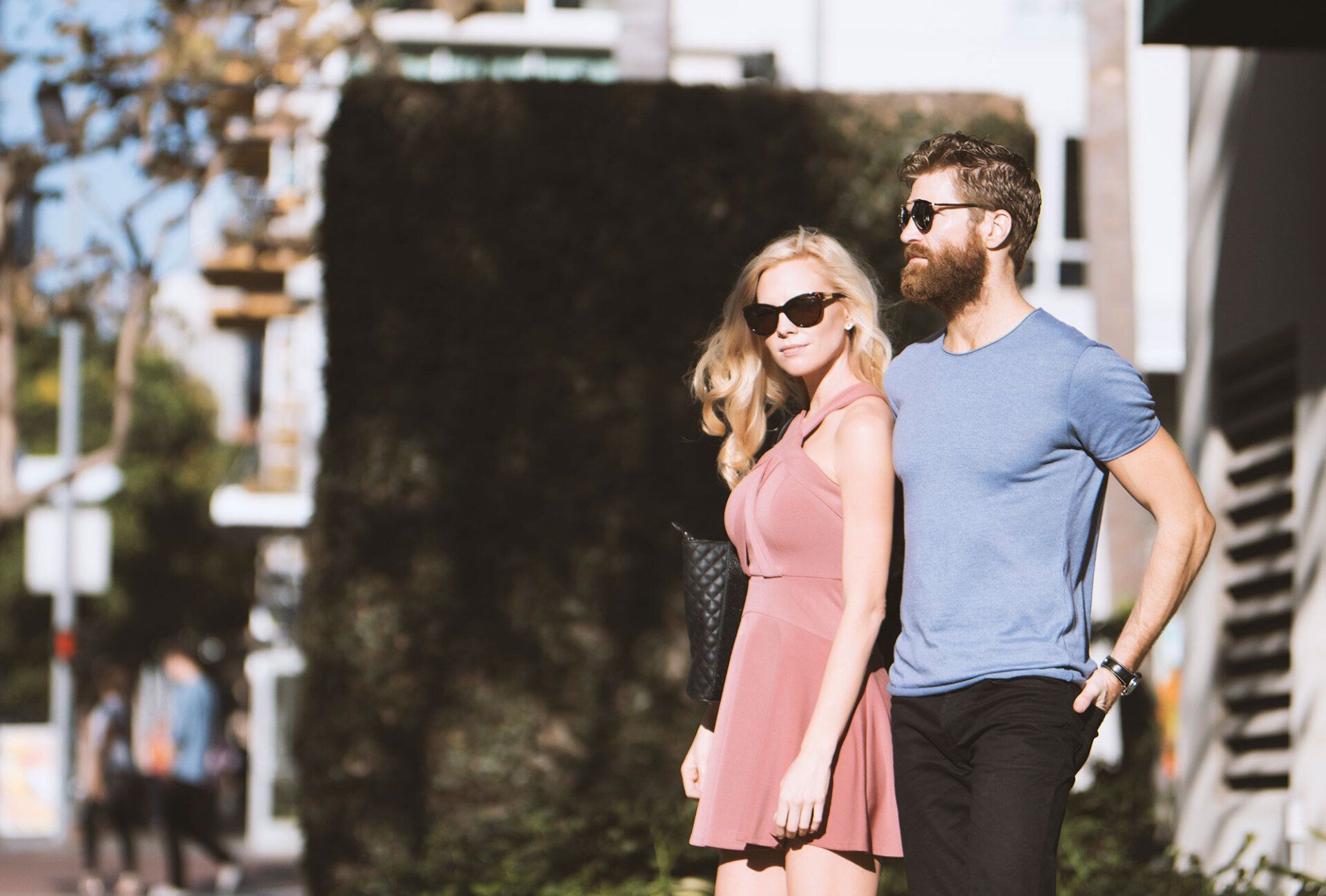 Woman in pink dress and man with beard walk together outside, both wearing sunglasses.
