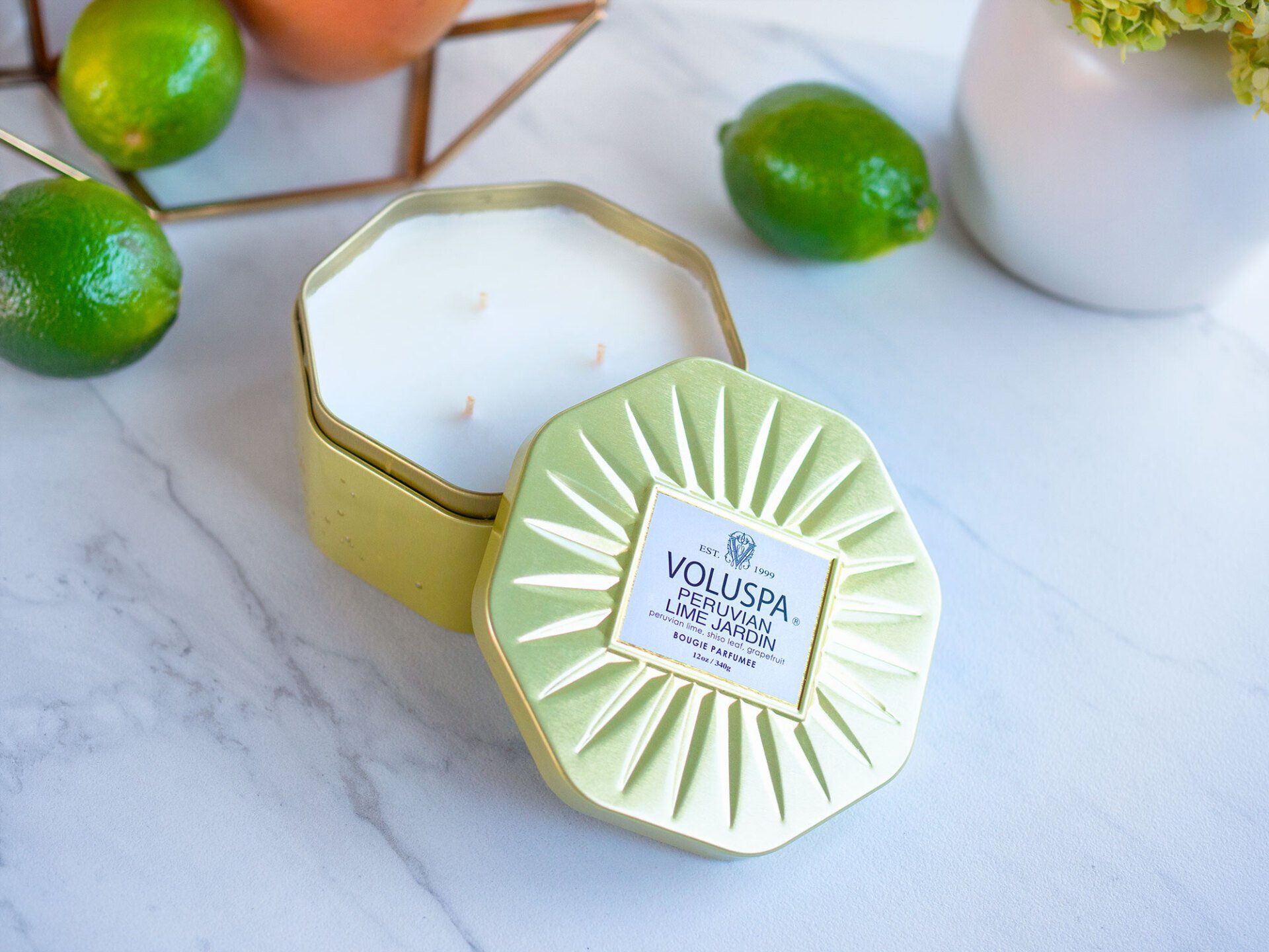 Gold hexagonal candle with lid, surrounded by limes and citrus fruits.