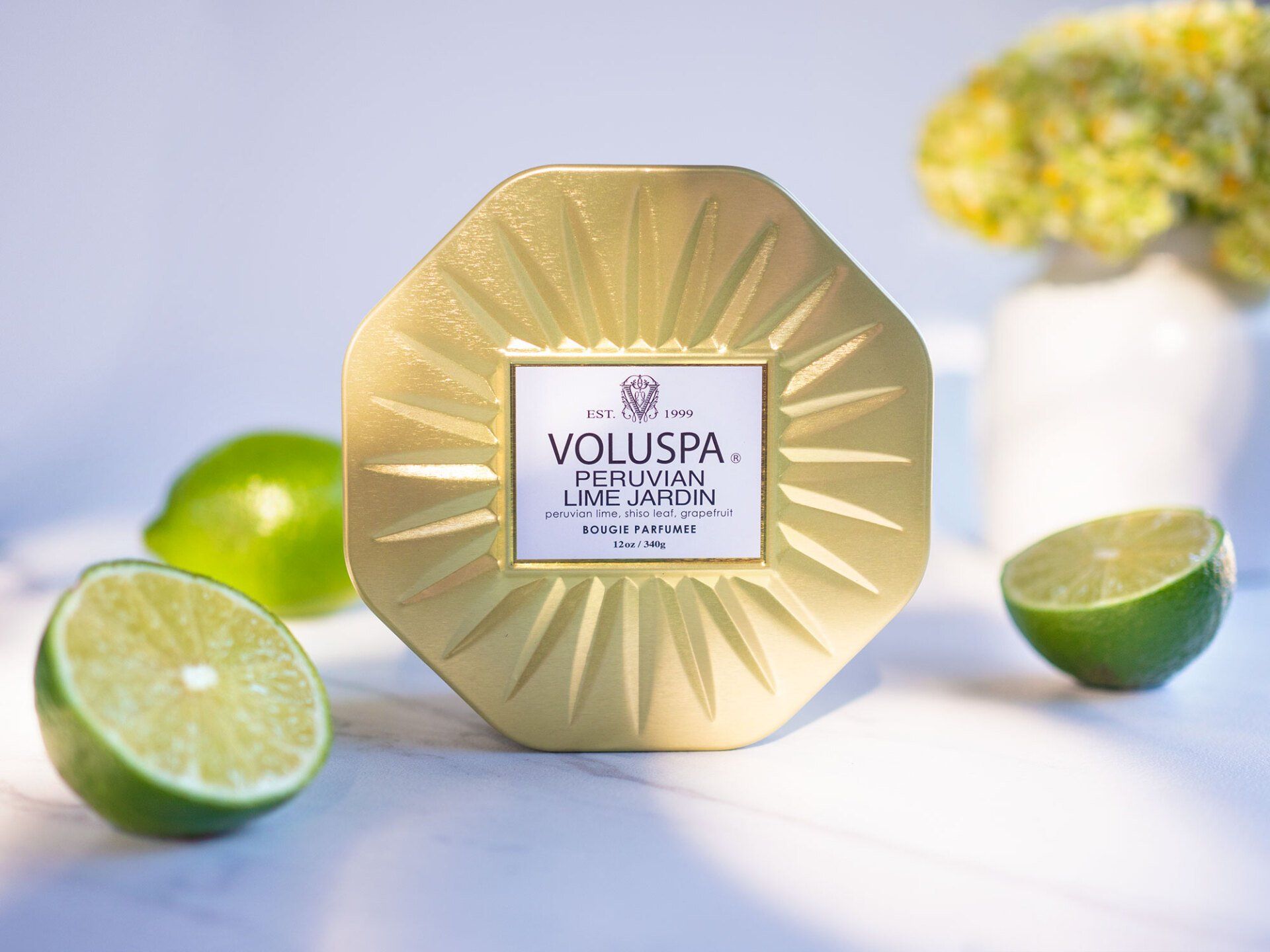 Gold candle in a decorative jar with limes on a marble surface, alongside a white vase with yellow flowers.