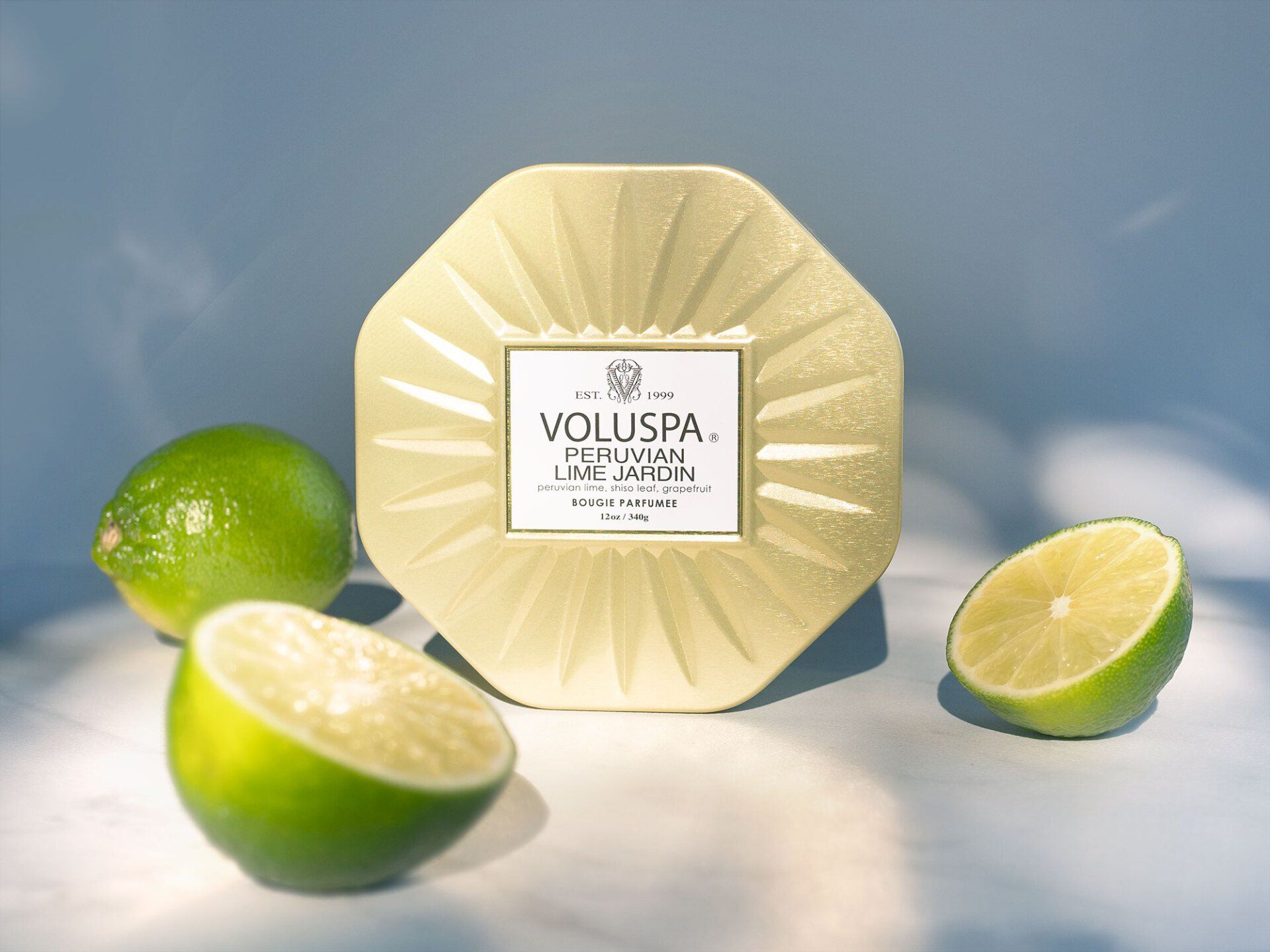 Gold Voluspa candle with limes, on light blue surface.