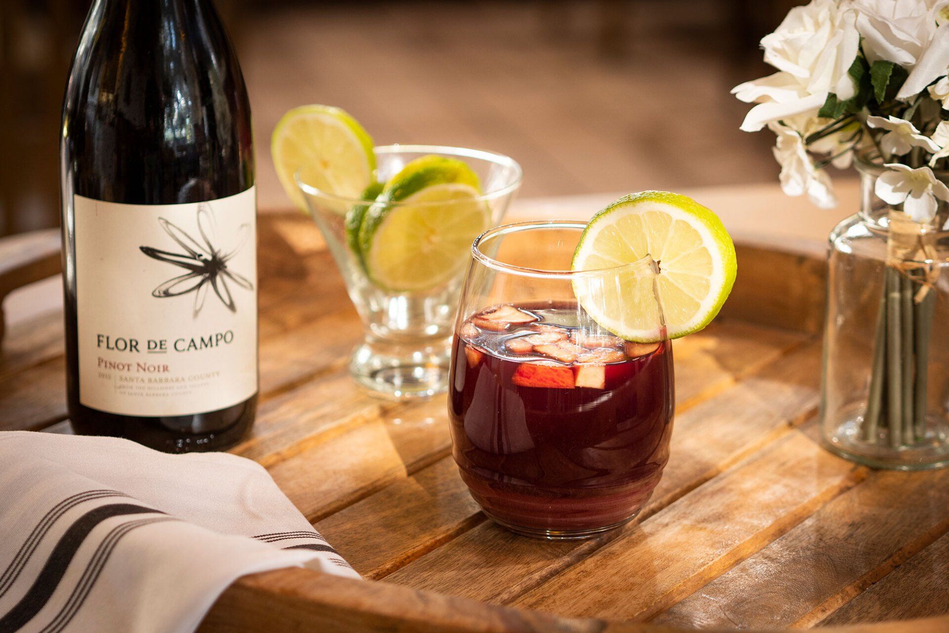 A glass of red sangria with lime and fruit, wine bottle, and flowers on a wooden tray.