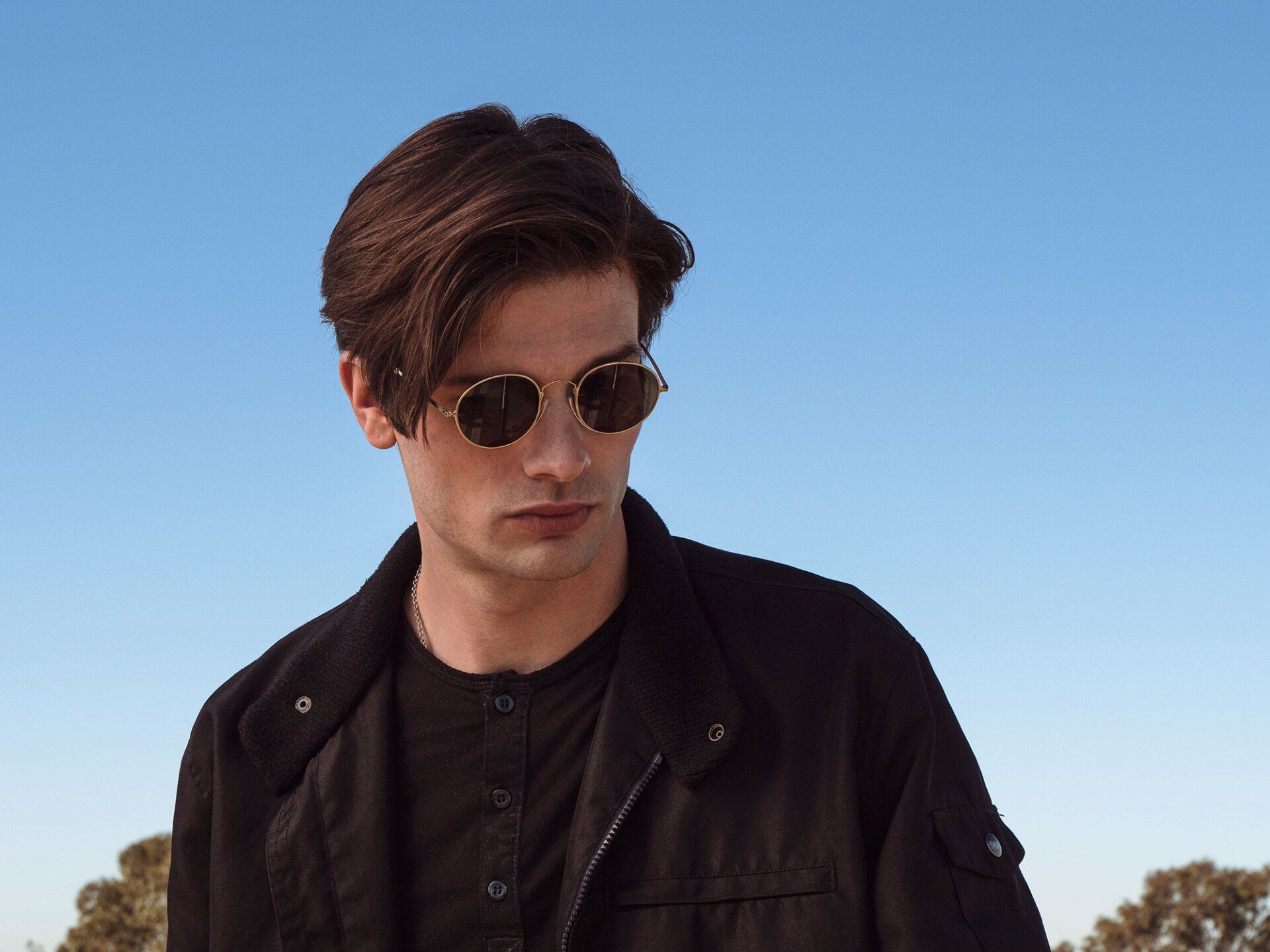 Man wearing sunglasses and a black jacket, looking down against a blue sky.