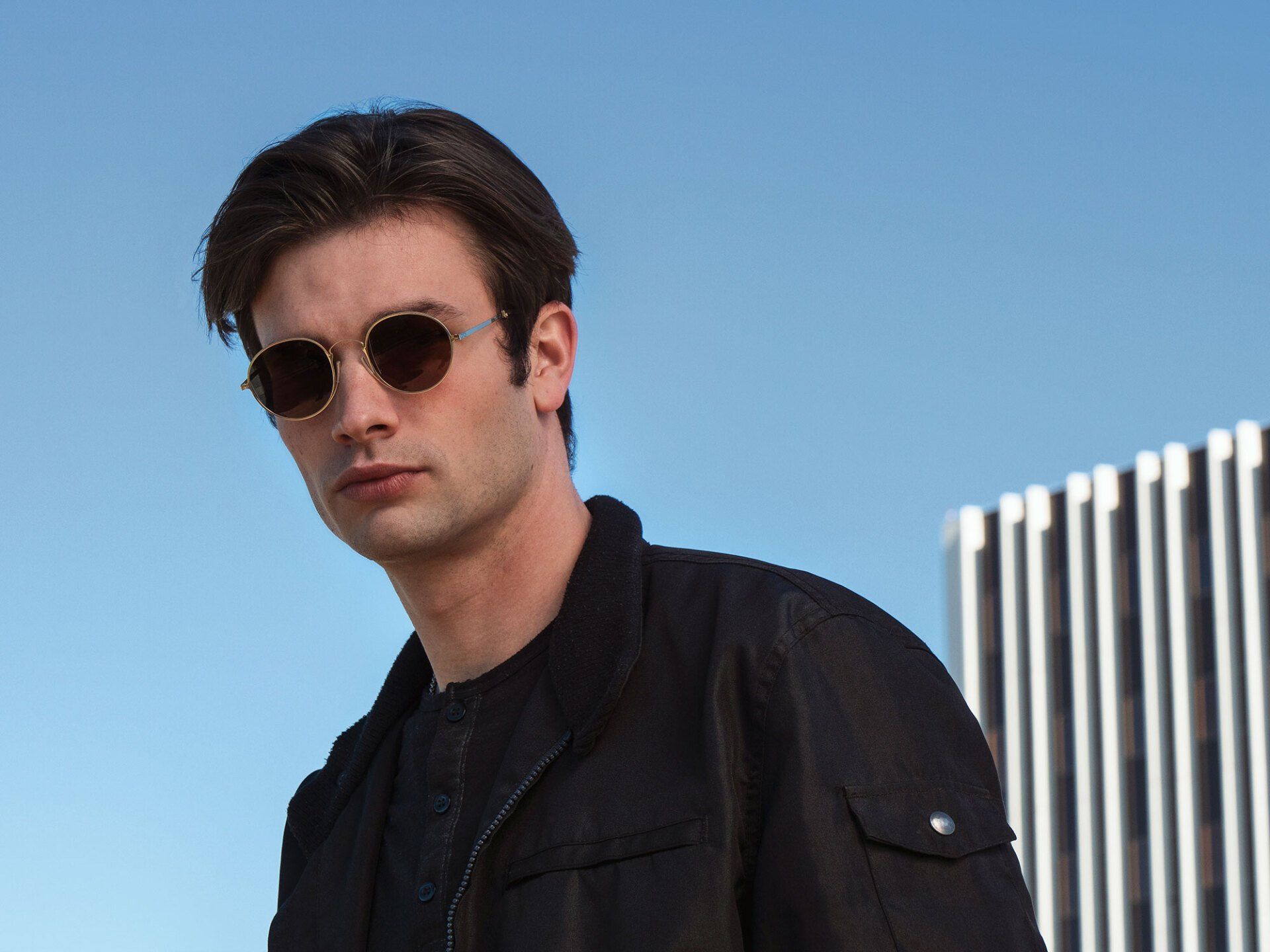 Man in black jacket and sunglasses, gazing at the camera, with a building and blue sky in background.