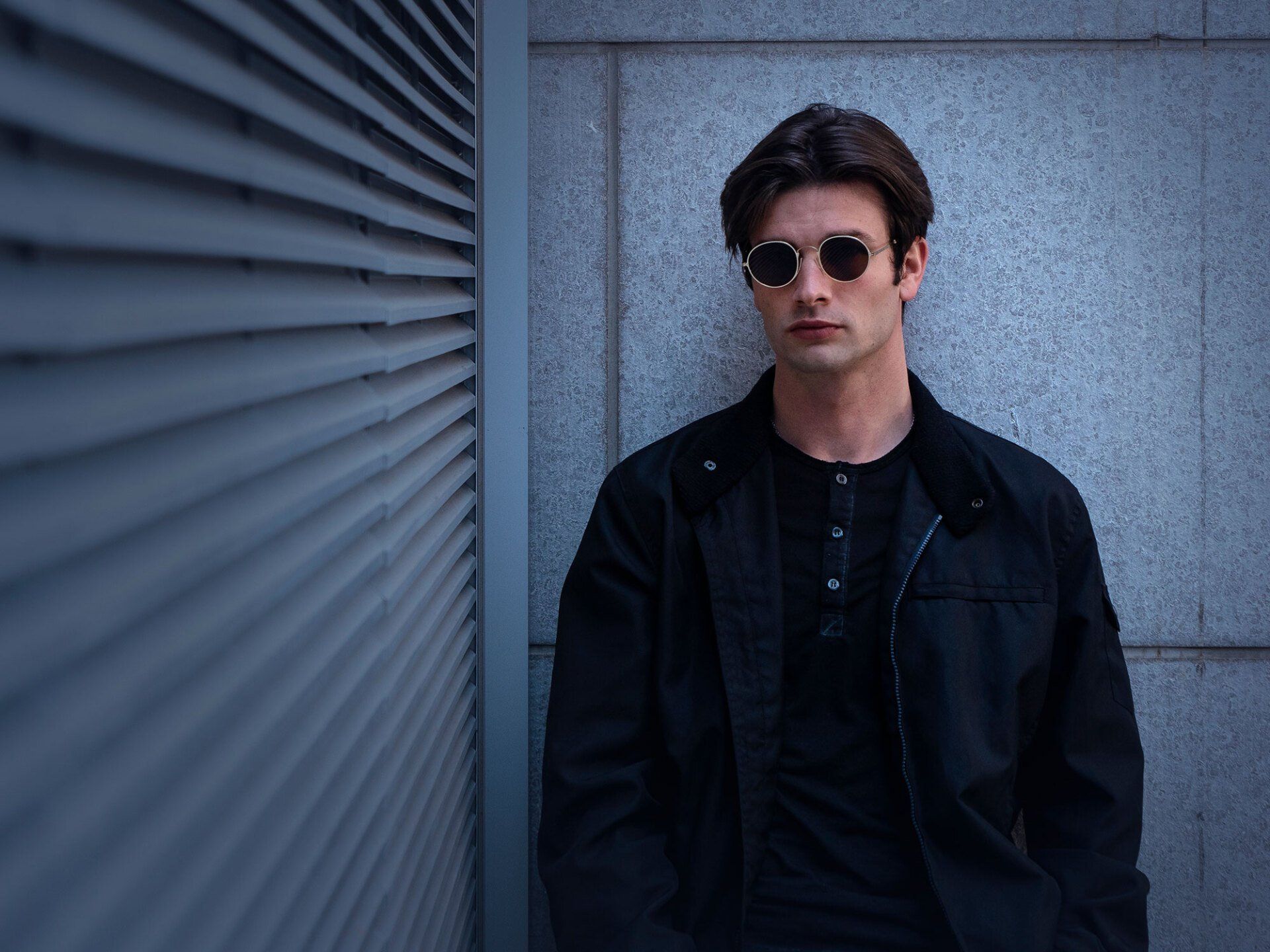 Man in sunglasses and black jacket leaning against a textured wall, looking forward.