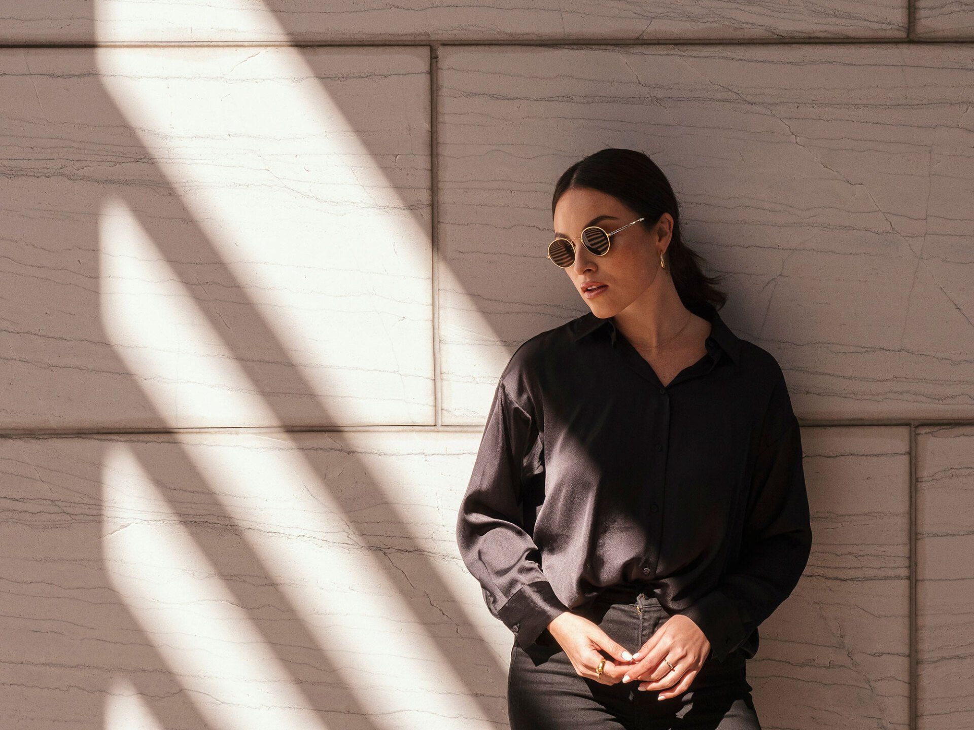 Woman in black clothing and sunglasses, leaning against a sunlit, textured wall.