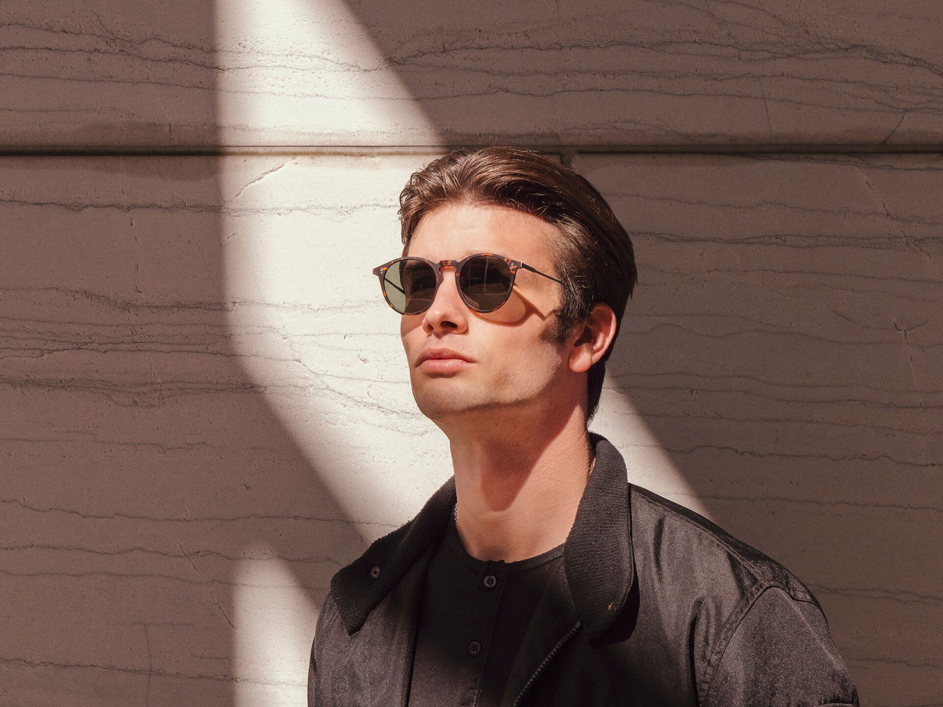 Man wearing sunglasses, looking upwards, in front of a marble wall with a patch of sunlight.