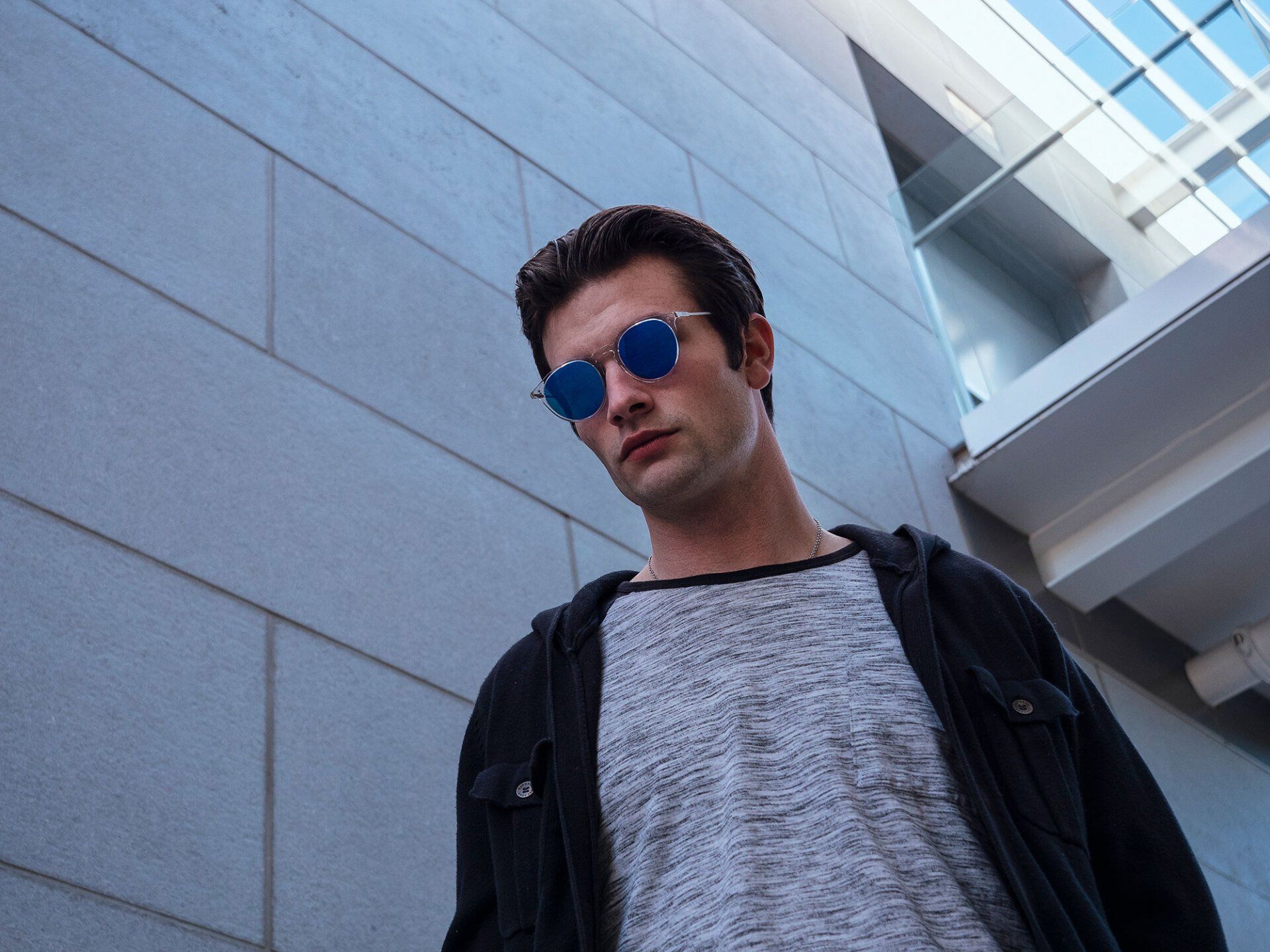 Man wearing blue sunglasses and a black jacket, standing outside a building.