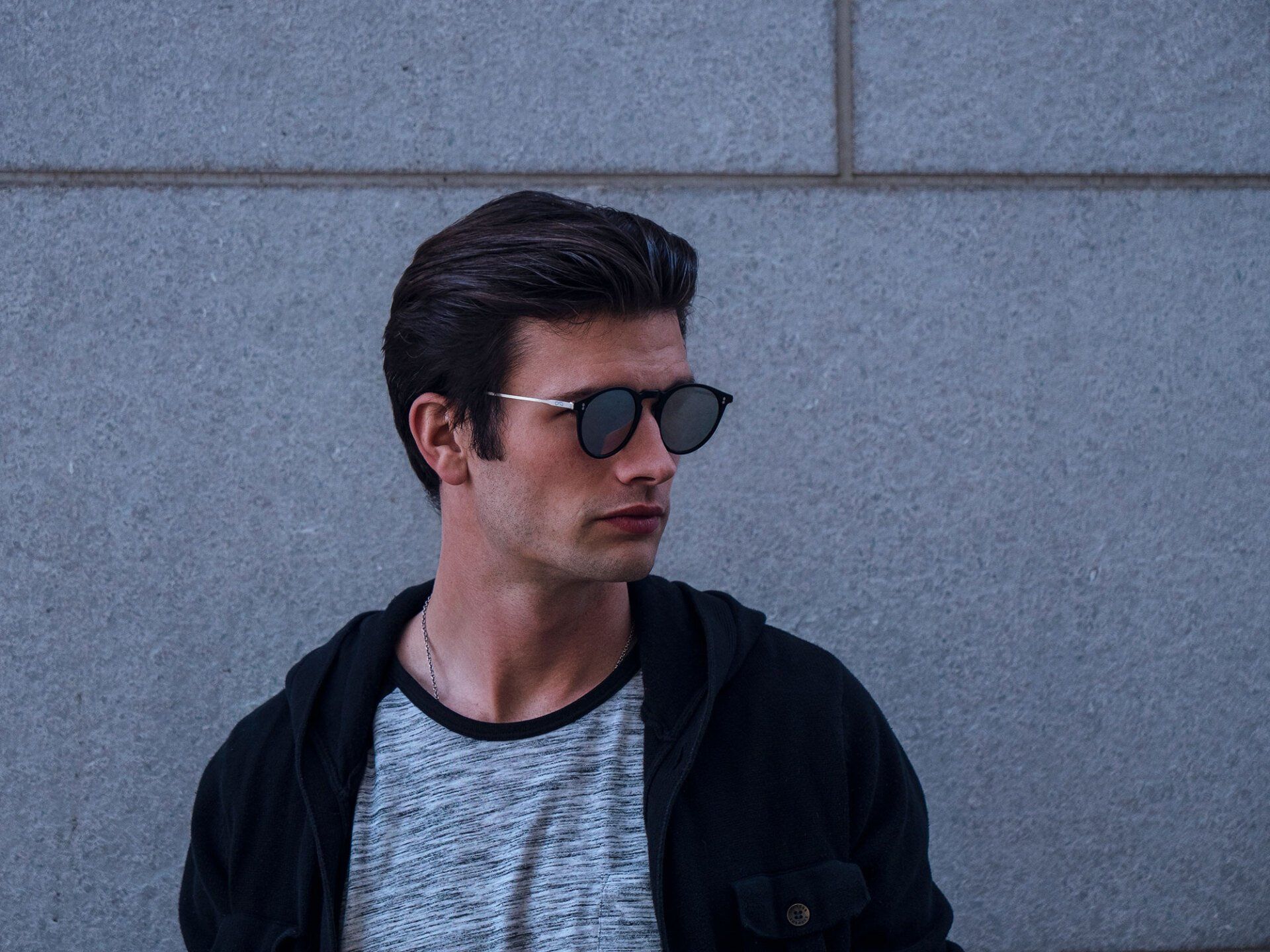 Man wearing sunglasses, looking left, in front of a gray wall. He wears a hoodie and t-shirt.