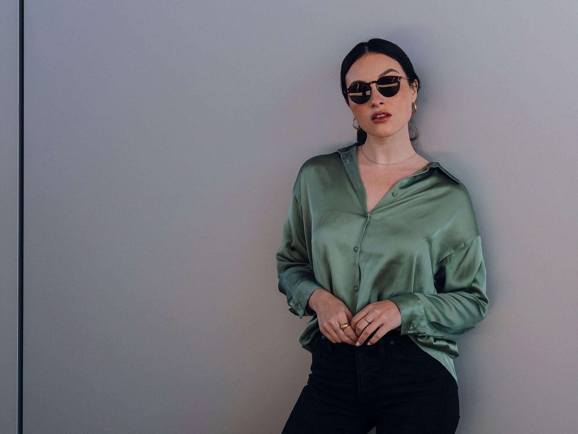 Woman in sunglasses and a green satin shirt, leaning against a gray wall.