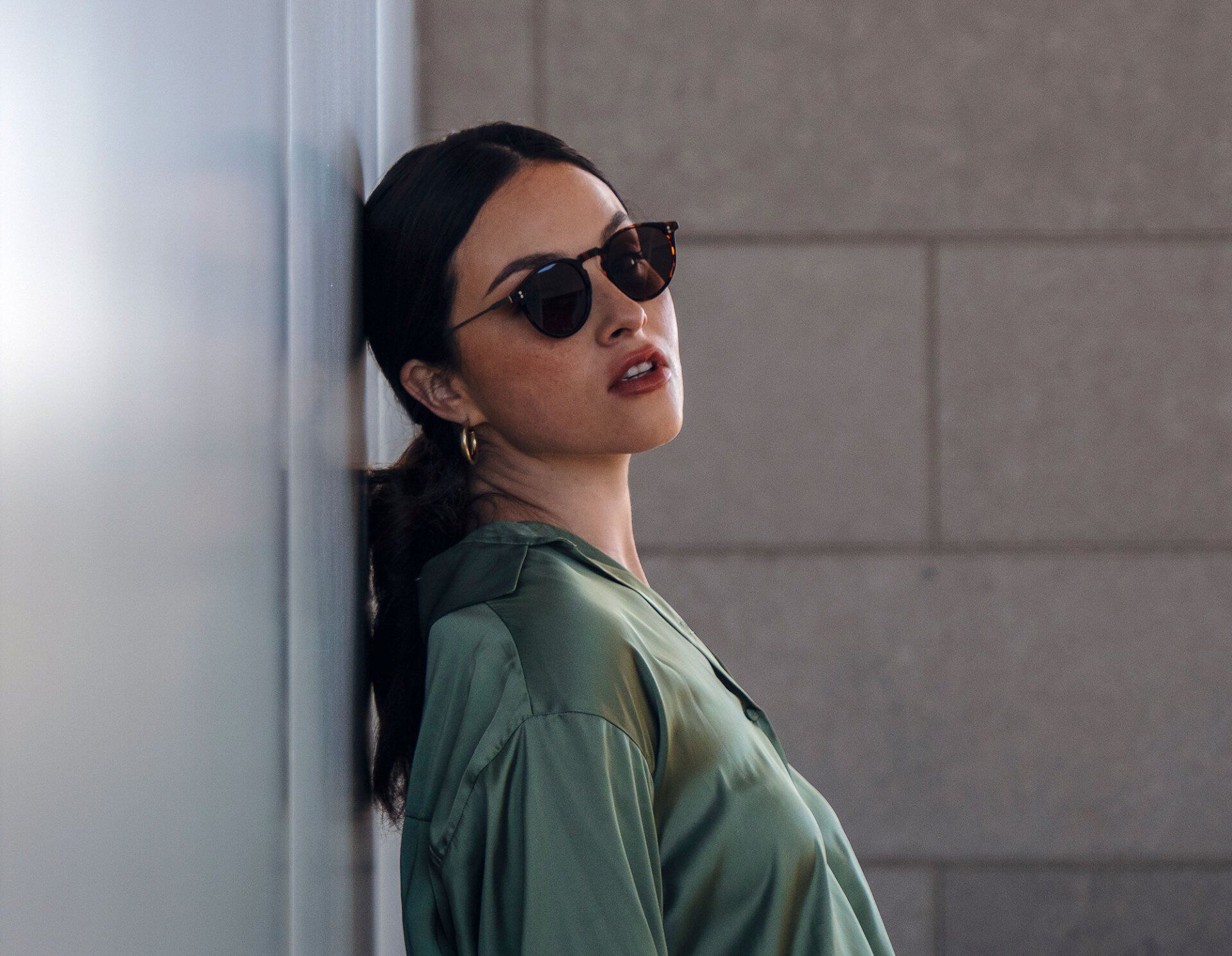 Woman in sunglasses leans against a wall, wearing a green blouse.