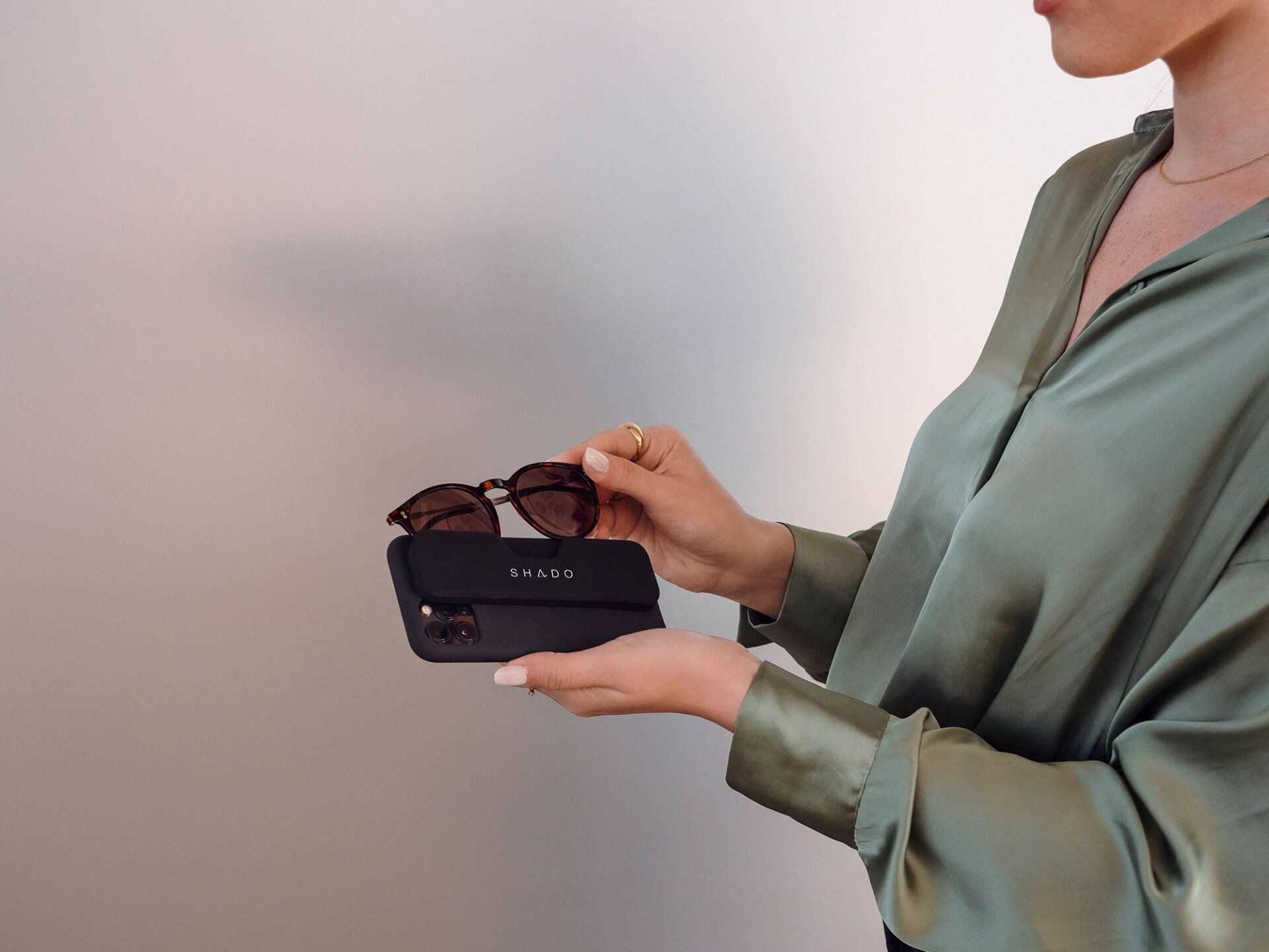 Woman holding sunglasses above a black case, wearing a green blouse against a white background.