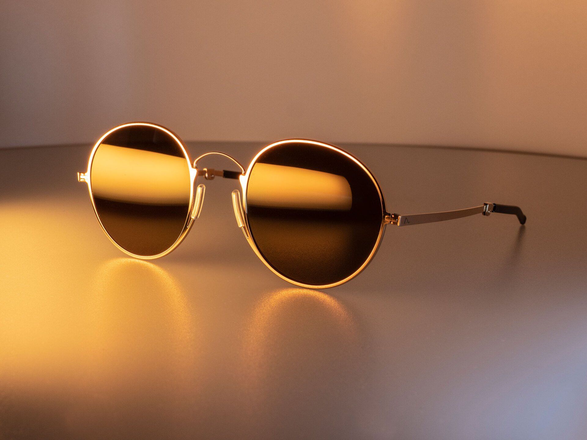Round gold-framed sunglasses with dark lenses on a reflective surface, lit by warm light.
