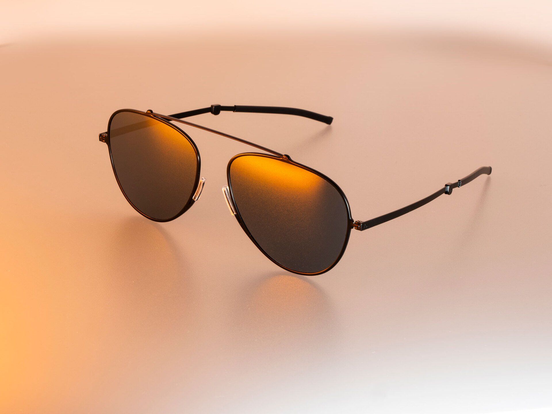 Black aviator sunglasses with orange mirrored lenses on a light orange surface.