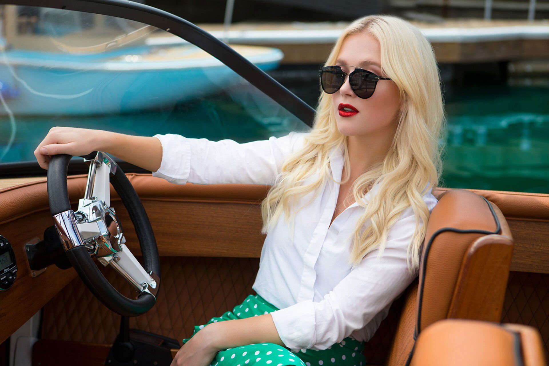 Blonde woman wearing sunglasses drives a boat in a marina, wearing a white shirt and green skirt.