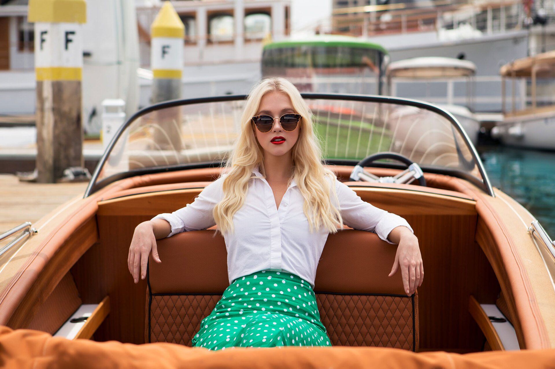 Woman in sunglasses sitting in a brown motorboat, wearing a white shirt and green skirt at a marina.