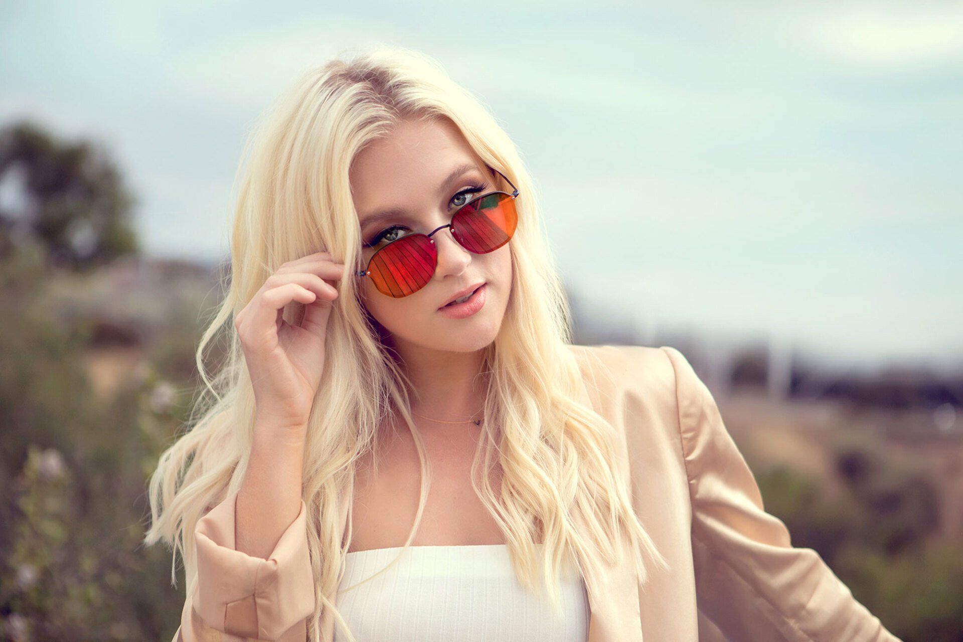 Woman with blonde hair wearing red sunglasses and tan blazer outdoors, looking at camera.