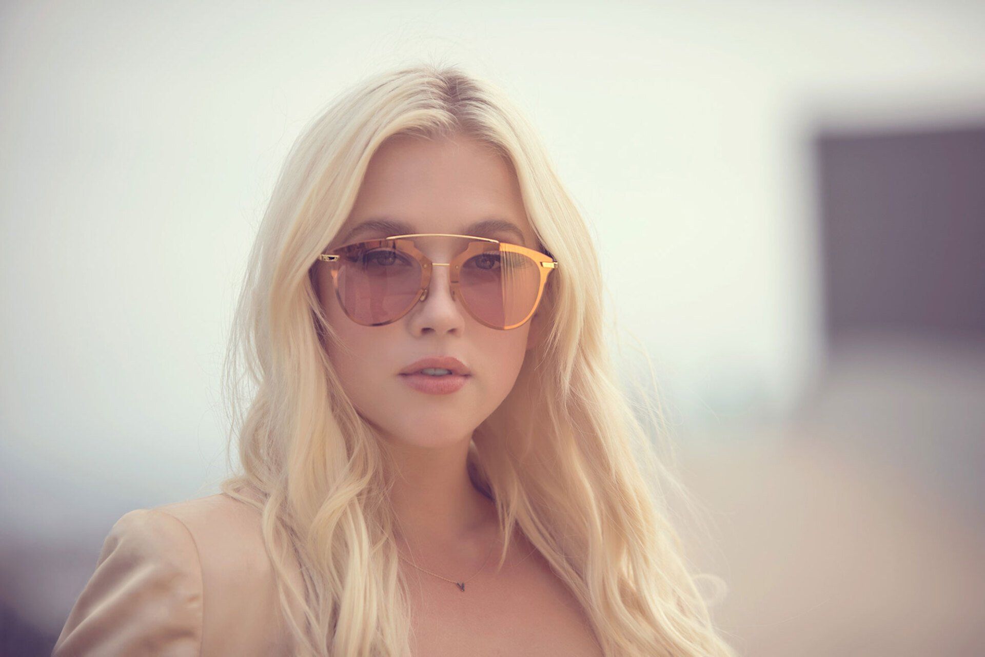 Blonde woman wearing sunglasses, a blazer, with blurred building background.