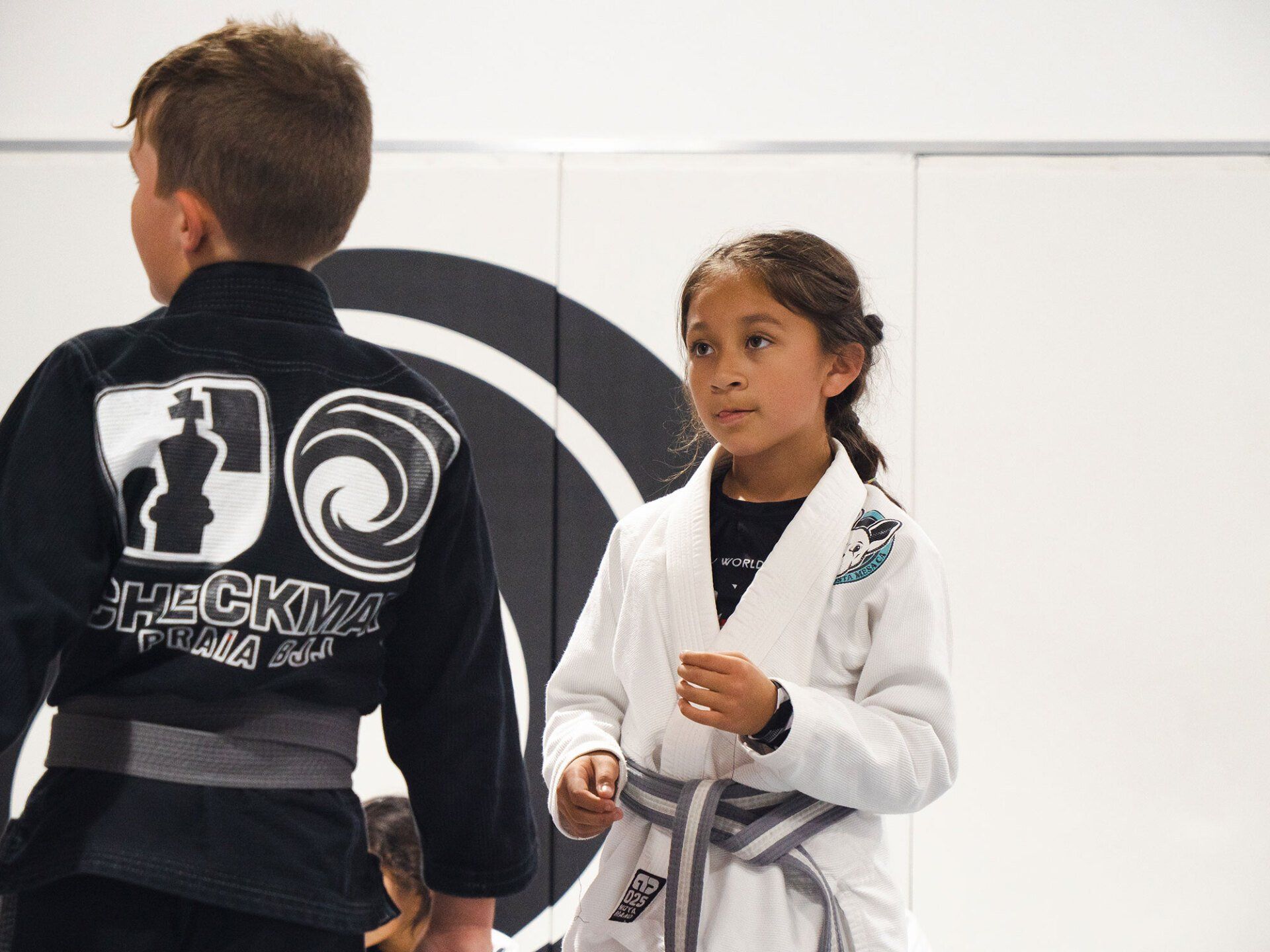 Girl in a white gi and boy in black gi on a mat, possibly in a martial arts class.
