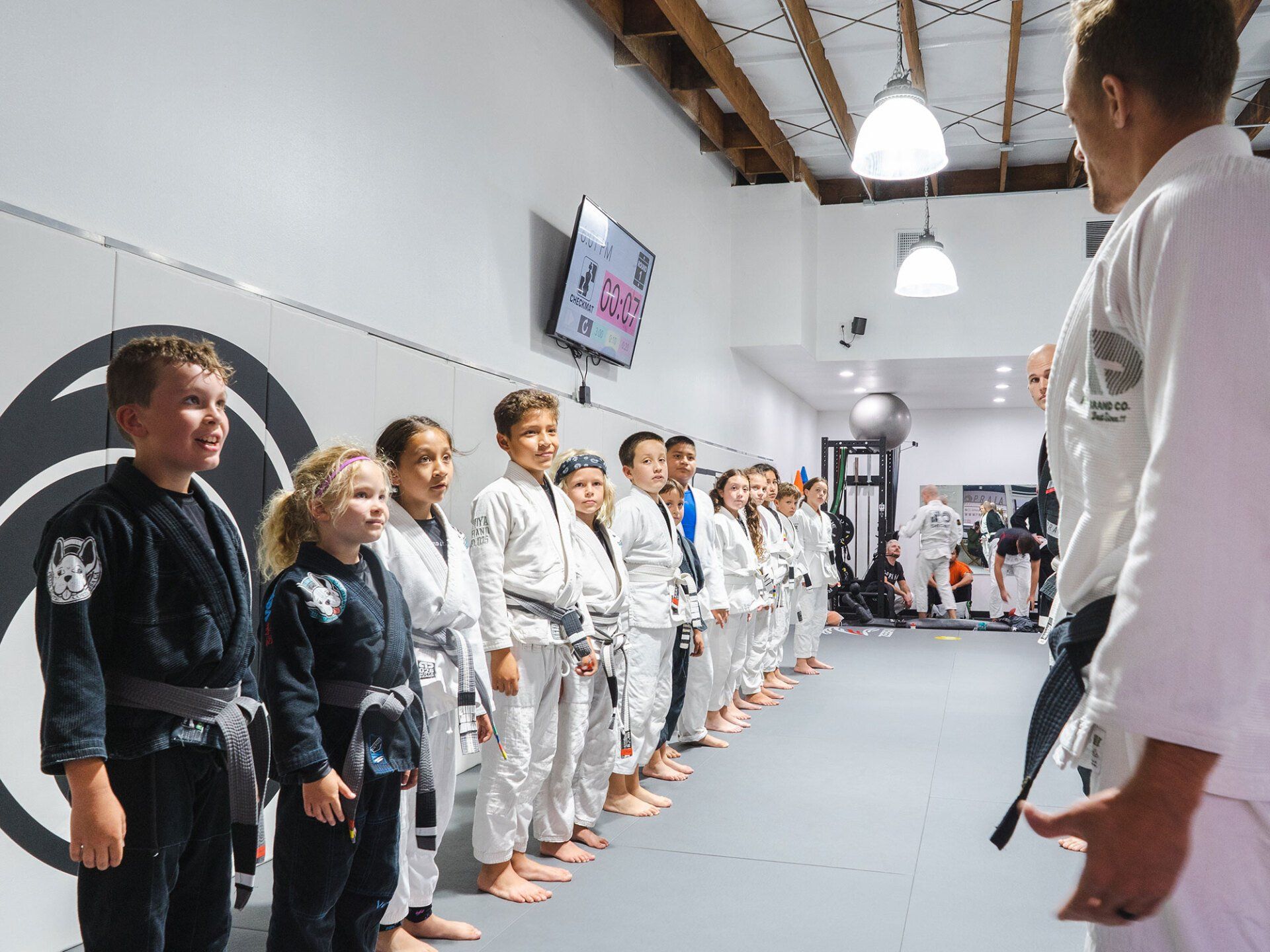 Children in white and black gis line up in a Jiu-Jitsu gym, facing a coach.