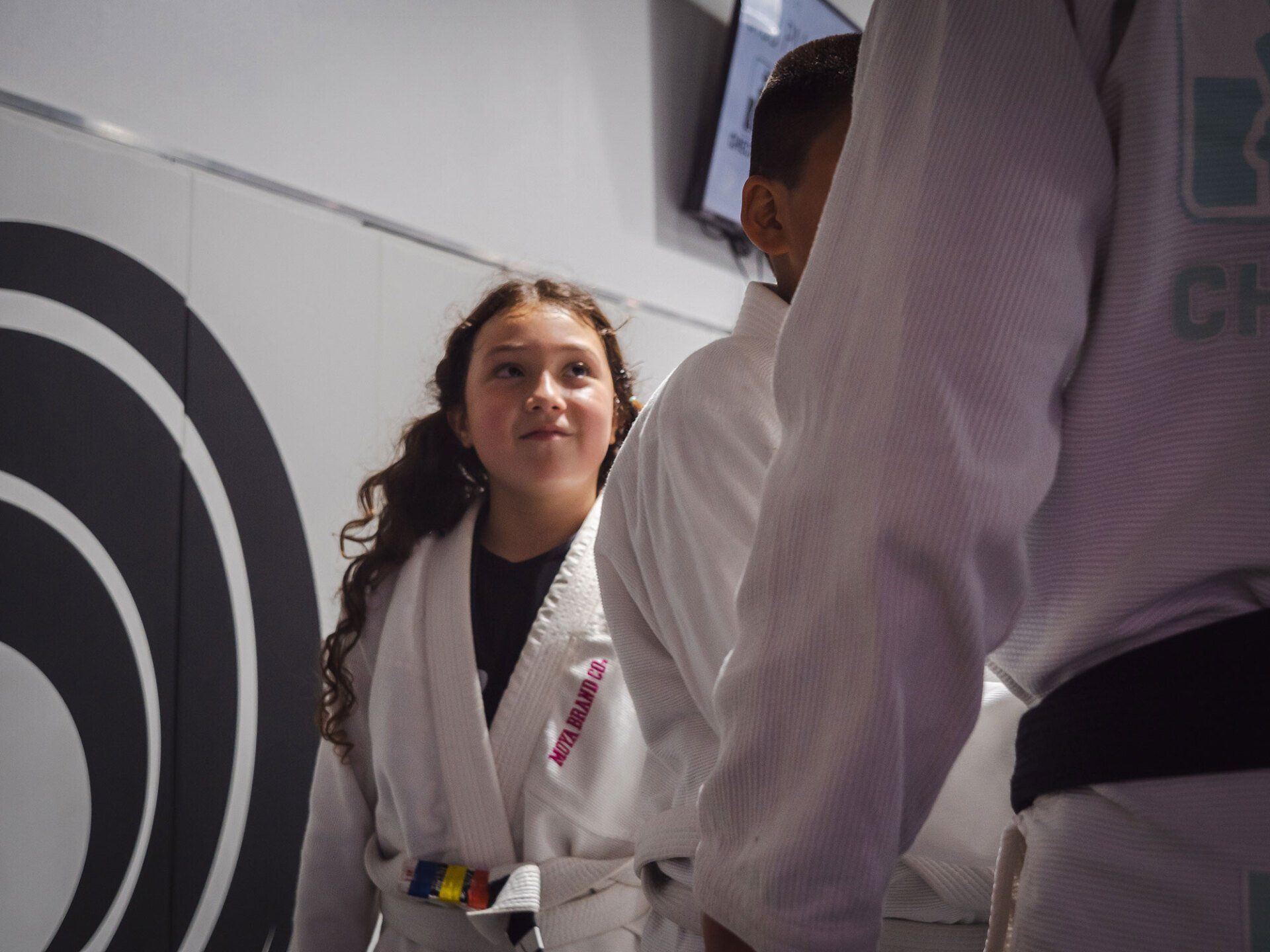 Young person in white martial arts uniform looking toward another person in a gym.