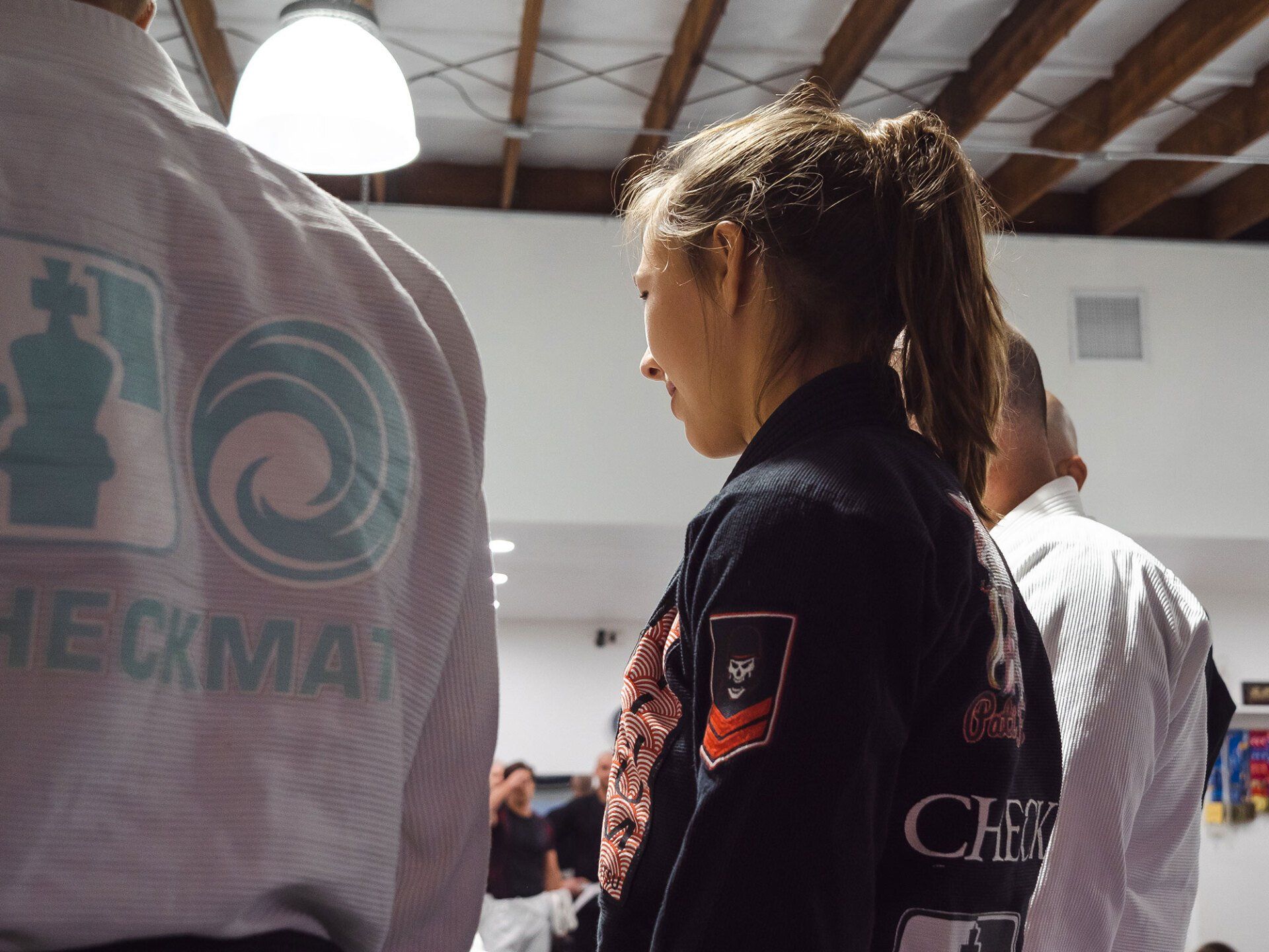 Woman in dark gi stands with others wearing light gis, Checmat logos visible.