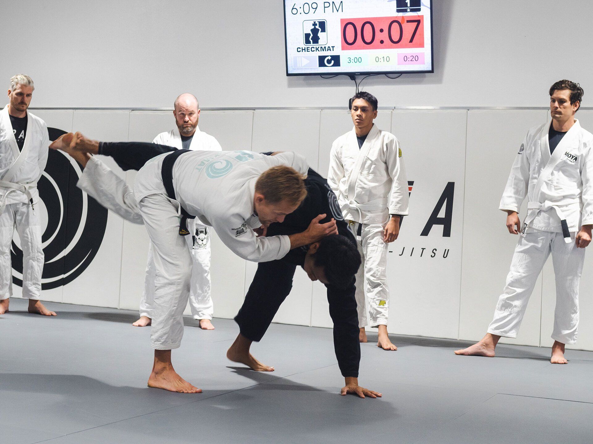Two men grappling in a jiu-jitsu class; one is lifting the other up. Several others watch. Timer displays 0:07.