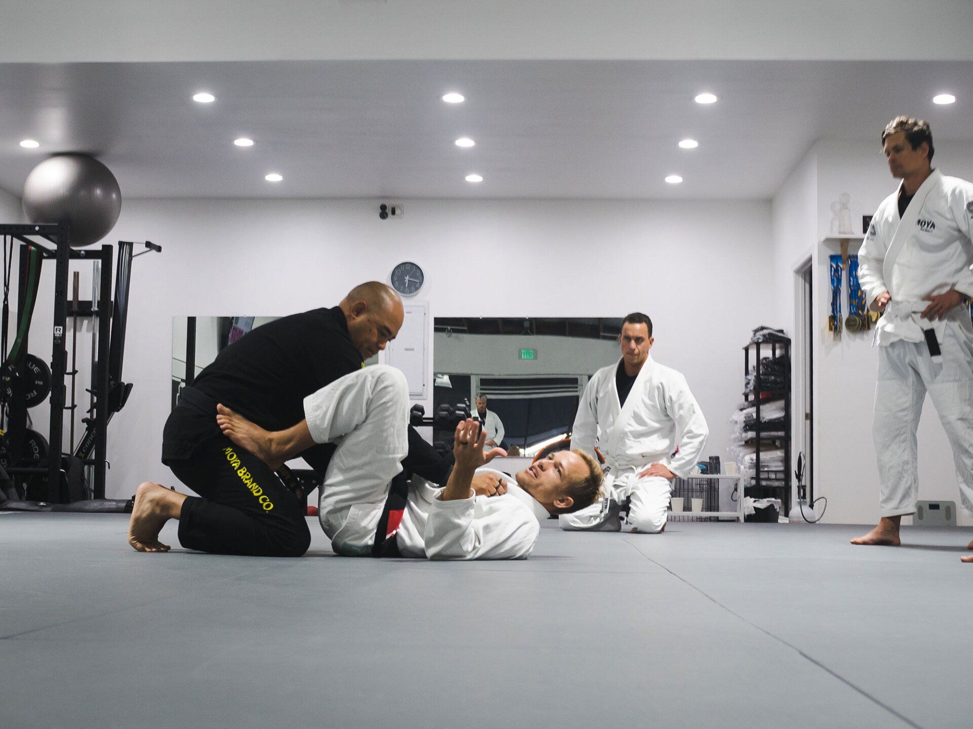 People practicing jiu-jitsu on a mat in a gym. One person is on their back being instructed.