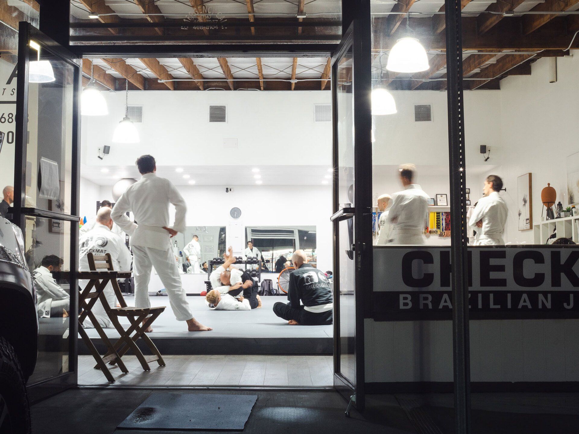 People in white gis inside a Brazilian Jiu-Jitsu gym with open doors at night.