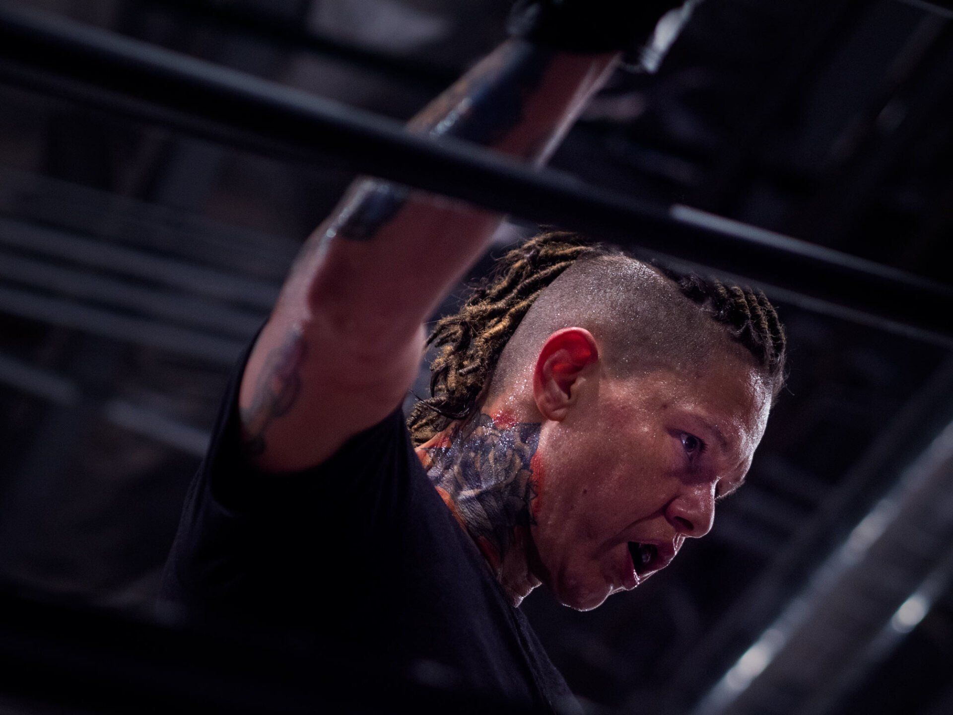 Wrestler, tattooed, with braided hair, raises arm in ring, looks strained.