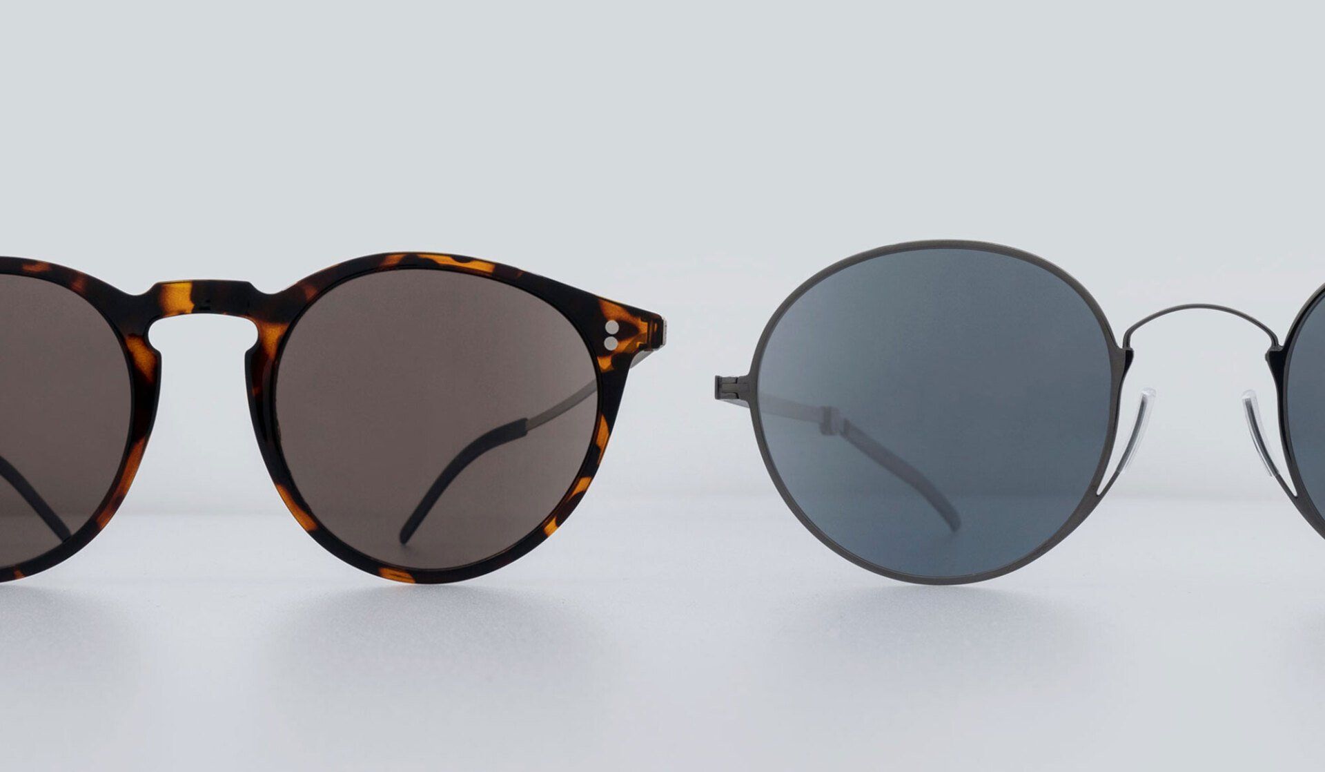 Two sunglasses on a white surface: tortoiseshell frames with brown lenses, and black frames with gray lenses.