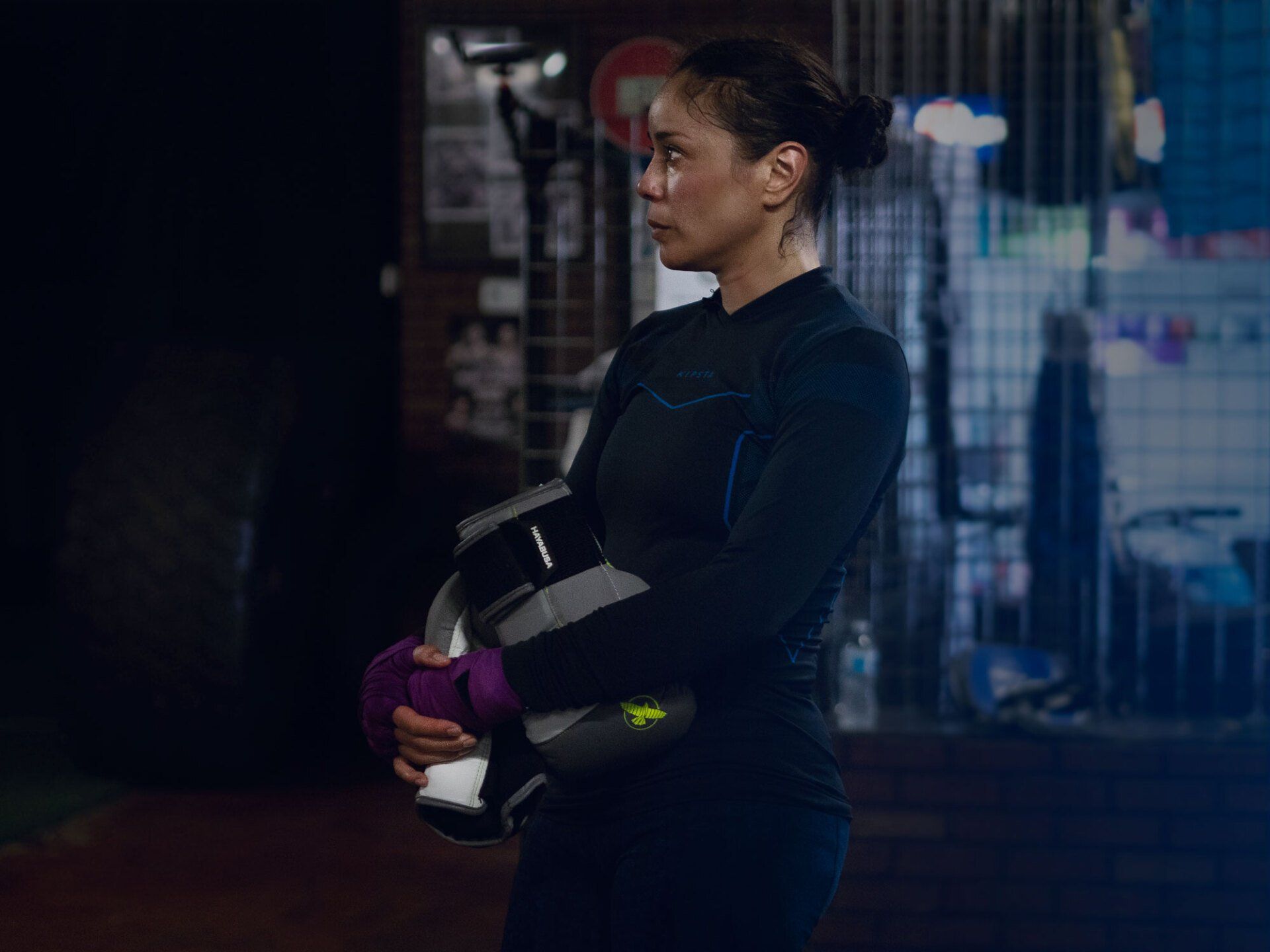 Woman in a boxing gym, holding gloves and looking away. Wearing a black athletic top, with purple hand wraps.