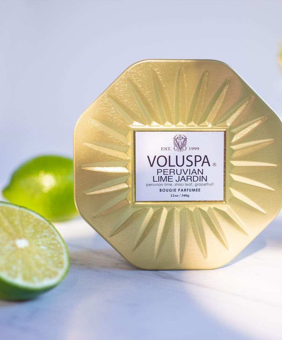 Gold Voluspa candle with label, surrounded by limes on a marble surface.