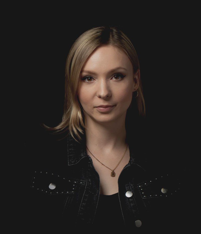 Woman with blonde hair wearing a black denim jacket, small necklace, looking at the camera. Dark background.