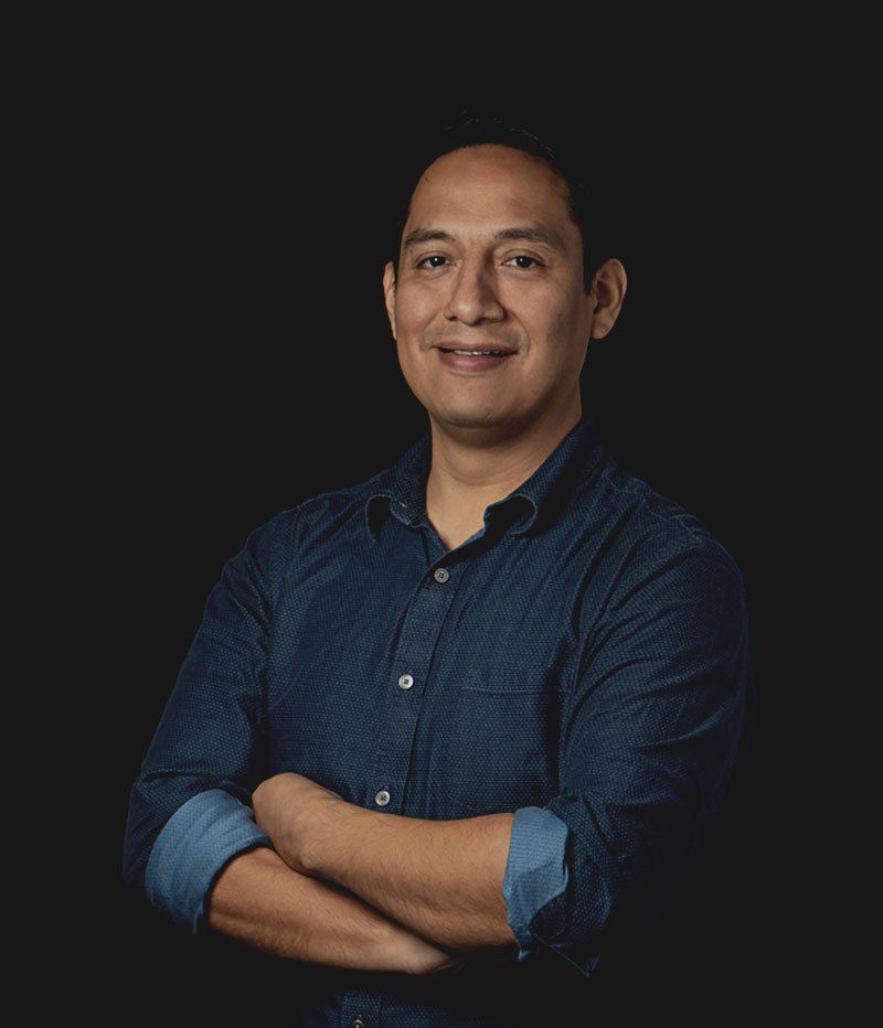 Man in a blue shirt with arms crossed, smiling, against a black background.