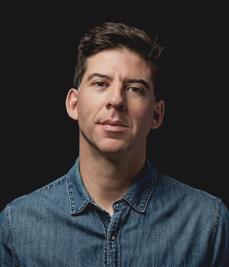 Man in denim shirt against a dark background, looking forward.