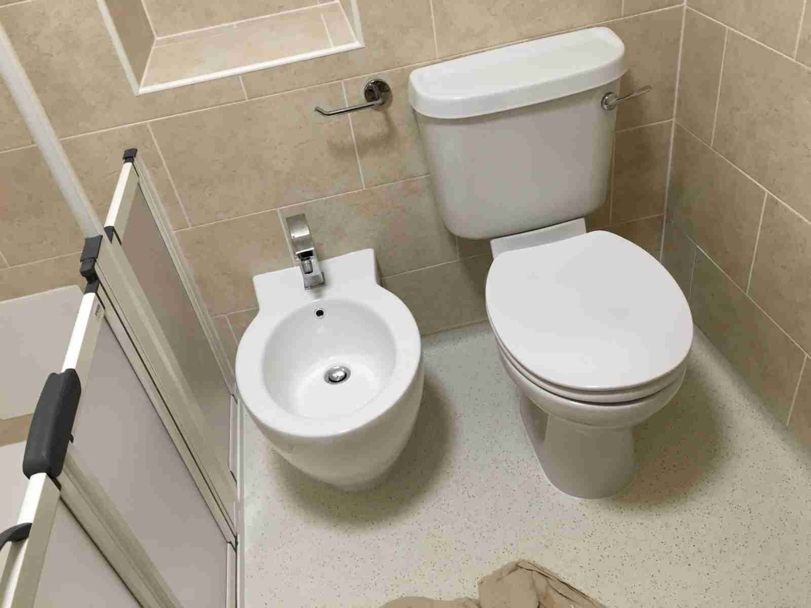 A white bidet and a matching white toilet sit side-by-side on a light-colored floor against beige tiled walls.