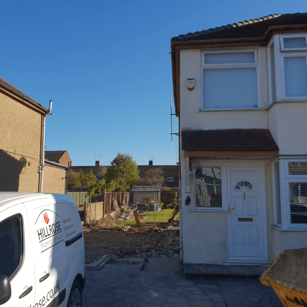 A semi-detached house with a skip in the driveway, beside a construction site with a white van parked in the foreground.