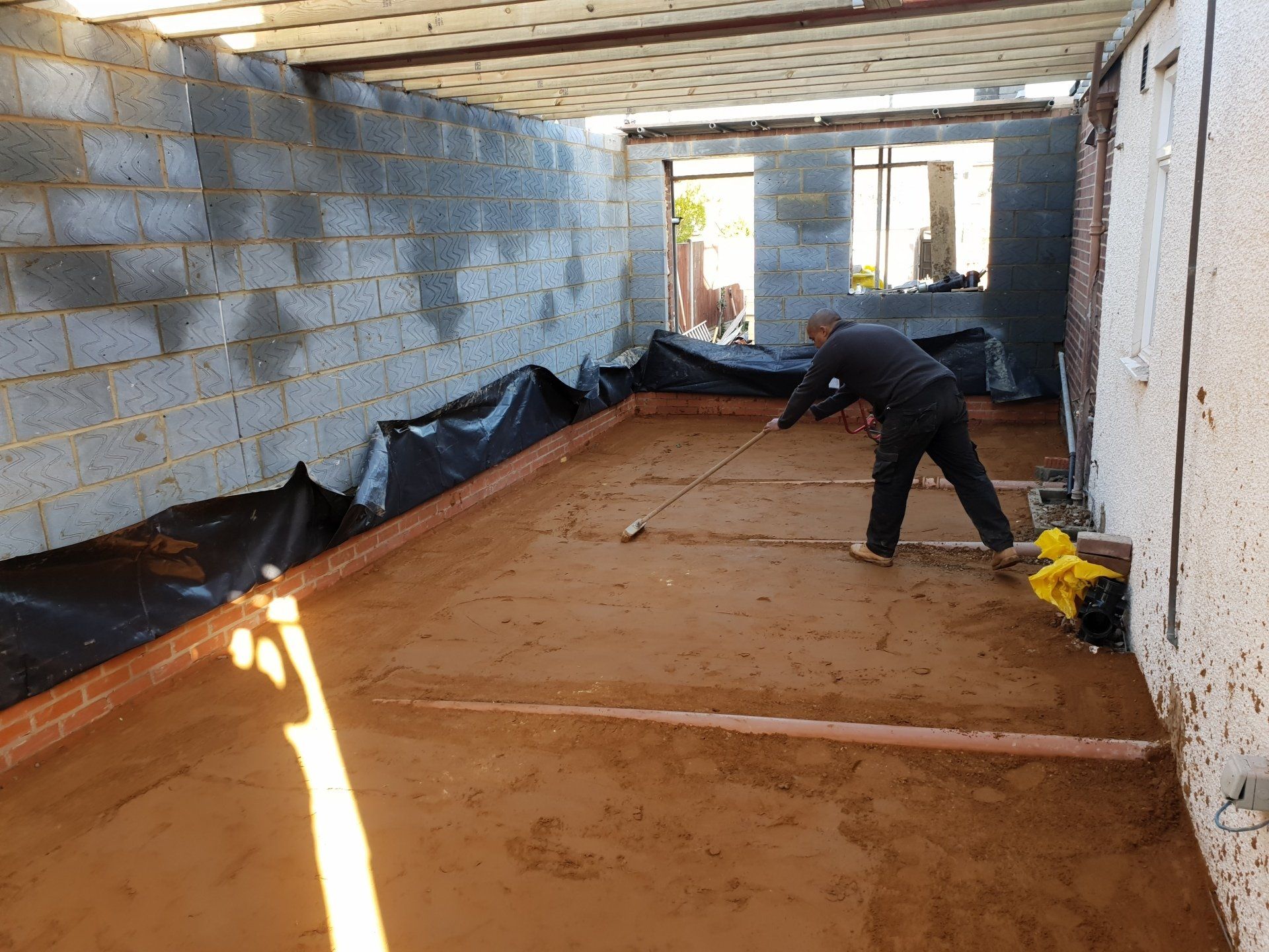 A worker uses a tool to level red sand across the floor of an indoor construction site with unfinished block walls.
