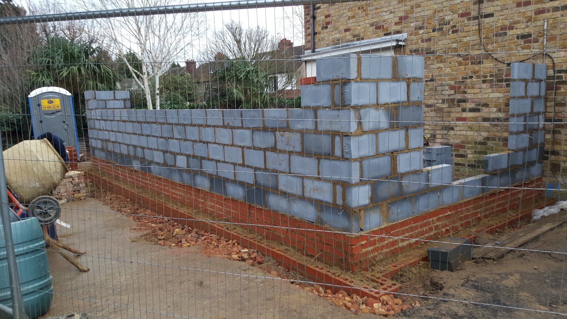 A construction site showing a low brick base with gray cinder block walls rising above it, viewed outdoors.