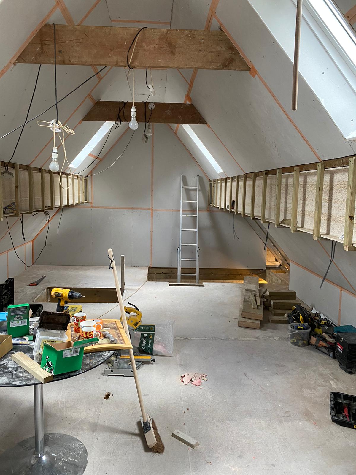 An attic under renovation with exposed wooden beams, unfinished drywall, a metal ladder, and scattered construction tools.