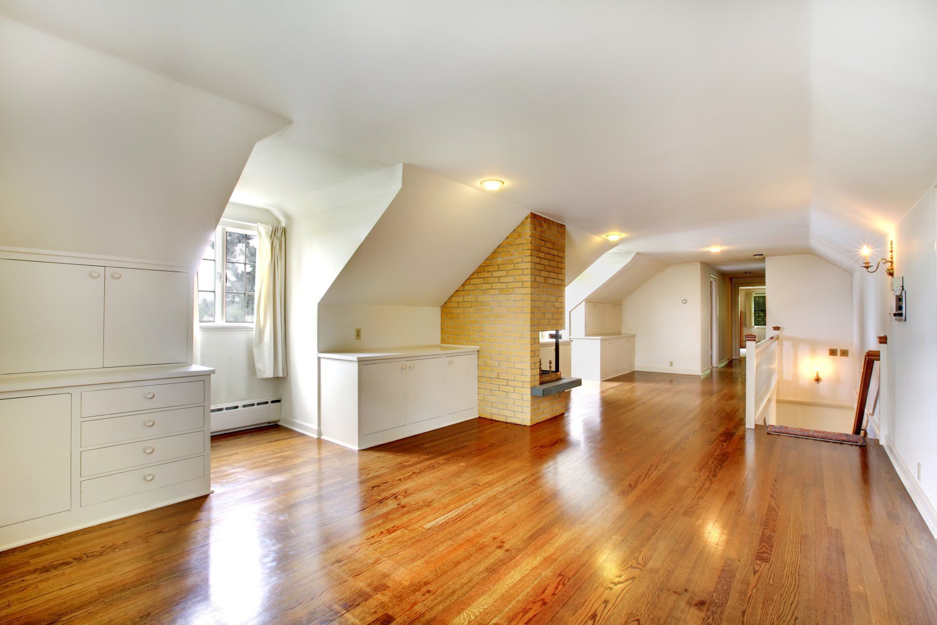 An empty, bright attic room with slanted white ceilings, hardwood floors, a brick fireplace, and built-in white cabinets.