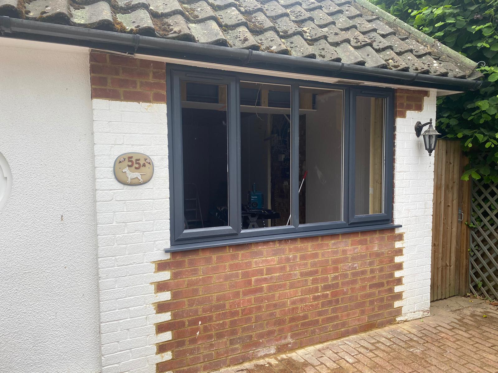 Exterior view of a home corner with white walls, red brick trim, and a modern grey window frame under a tiled roof.