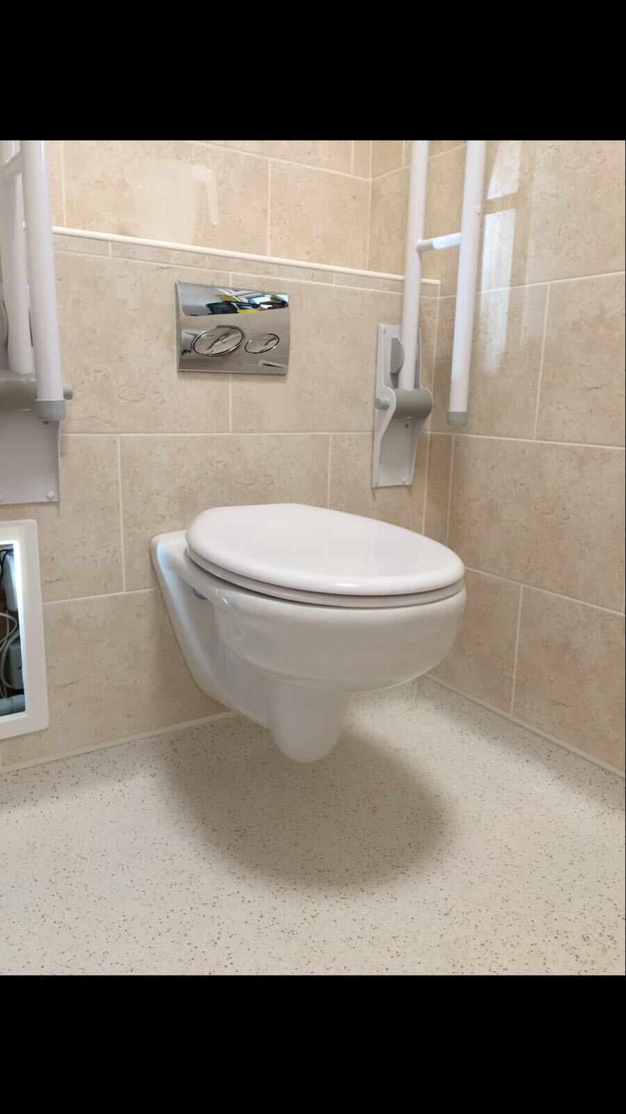 Wall-mounted toilet with a closed lid, grab rails, and flush plate in a tiled, beige accessible bathroom stall.